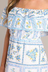 Close up view of this white & blue maxi dress featureing an elastic off-the-shoulder neckline an embroidered floral design, and a lined and tiered skirt.
