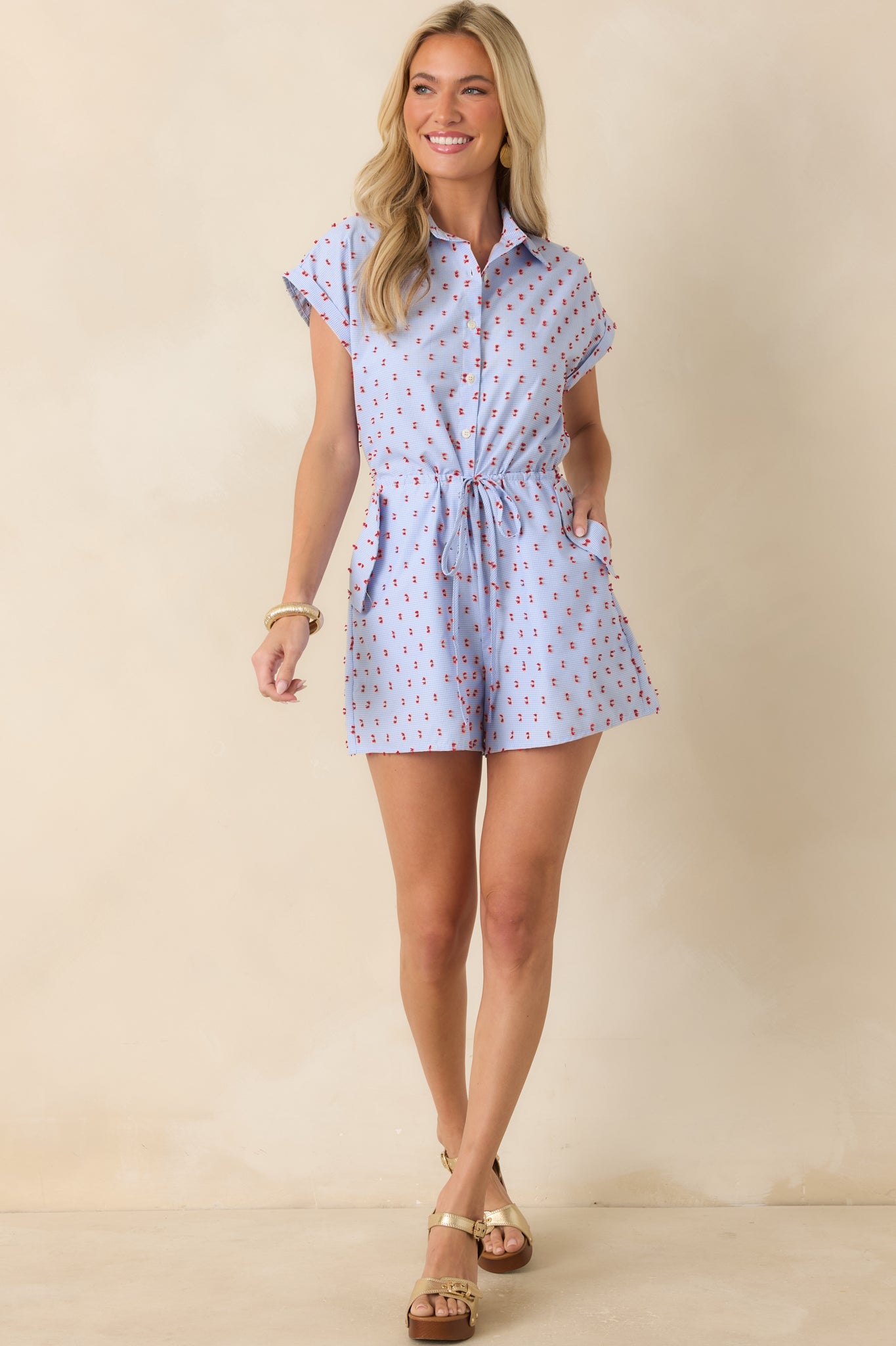 Blue gingham romper featuring red tufted embroidery, a collared neckline, and a relaxed shorts silhouette.