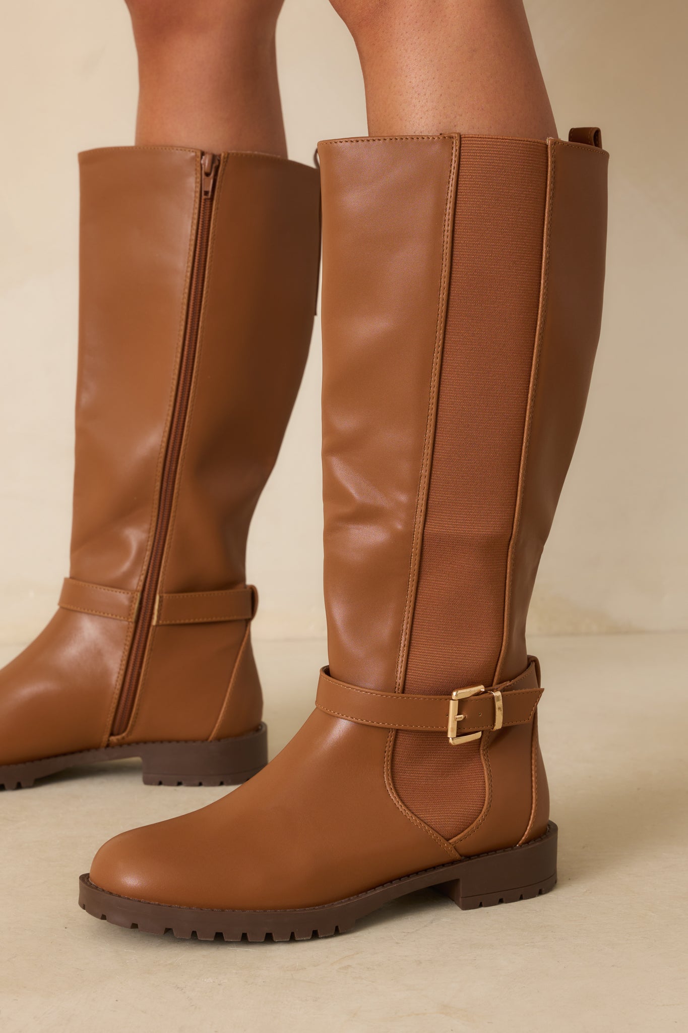 Side view of the caramel brown riding boots on beige, highlighting the elastic panels and ankle strap.