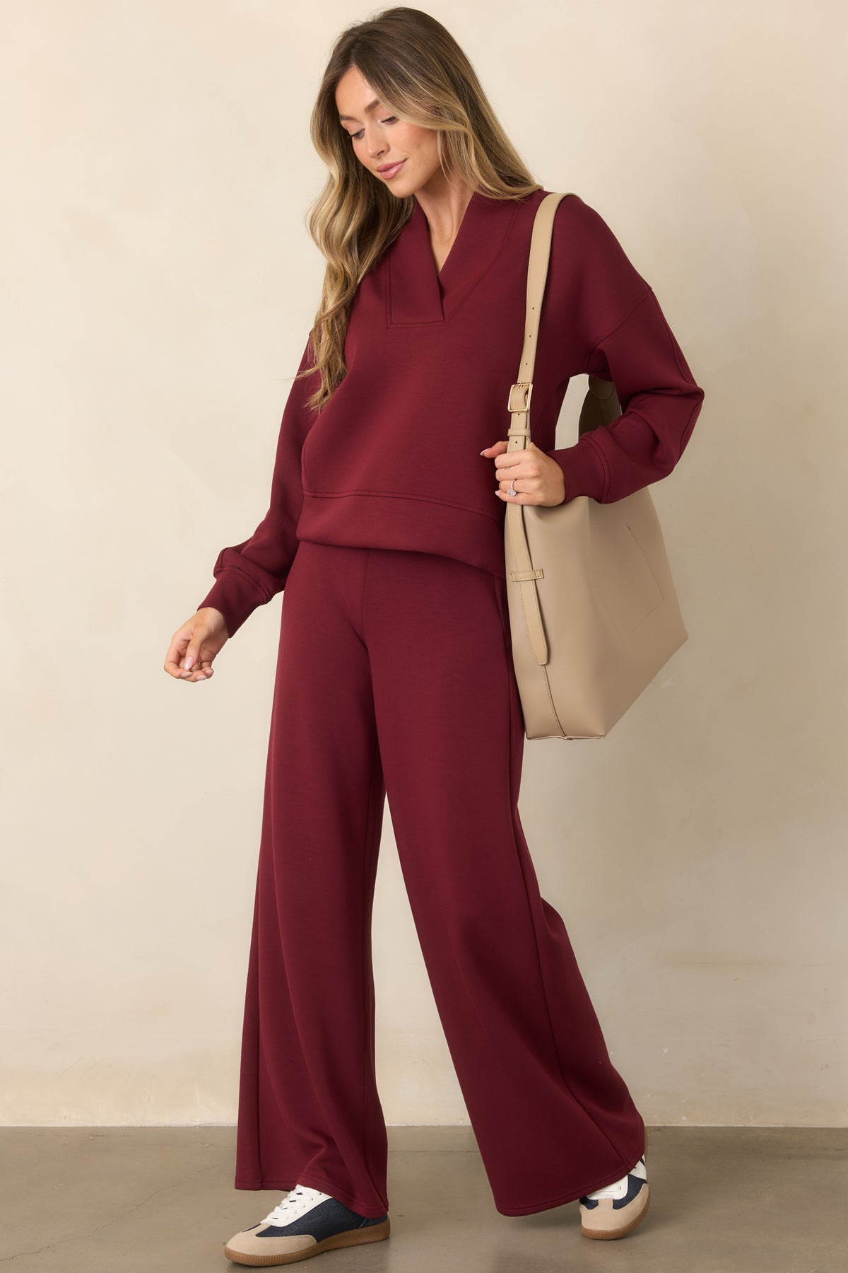 A woman modeling a matching burgundy lounge set consisting of a long-sleeve V-neck sweatshirt and wide-leg pants, styled with a beige tote bag and navy sneakers against a neutral beige studio background.
