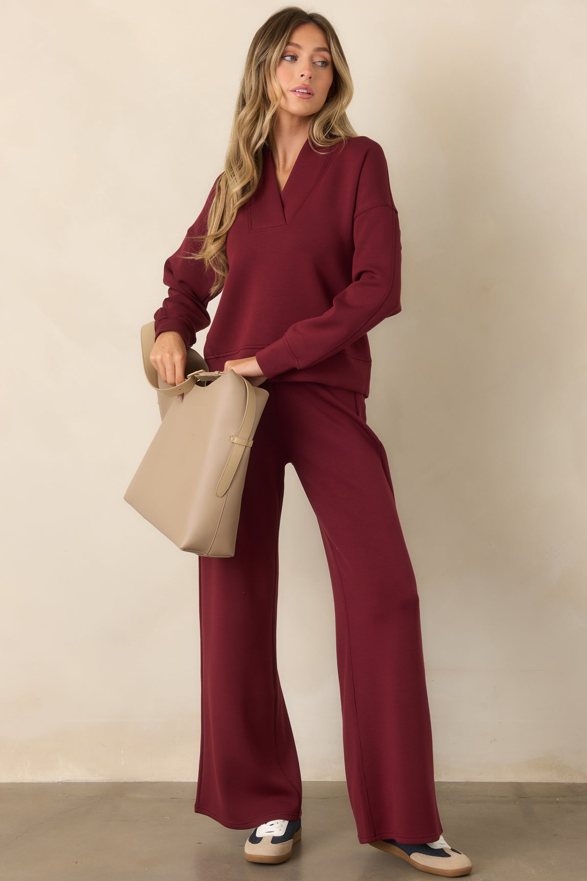 A woman modeling a matching burgundy long-sleeve V-neck top and wide-leg pants set, holding a beige tote bag against a neutral beige wall.