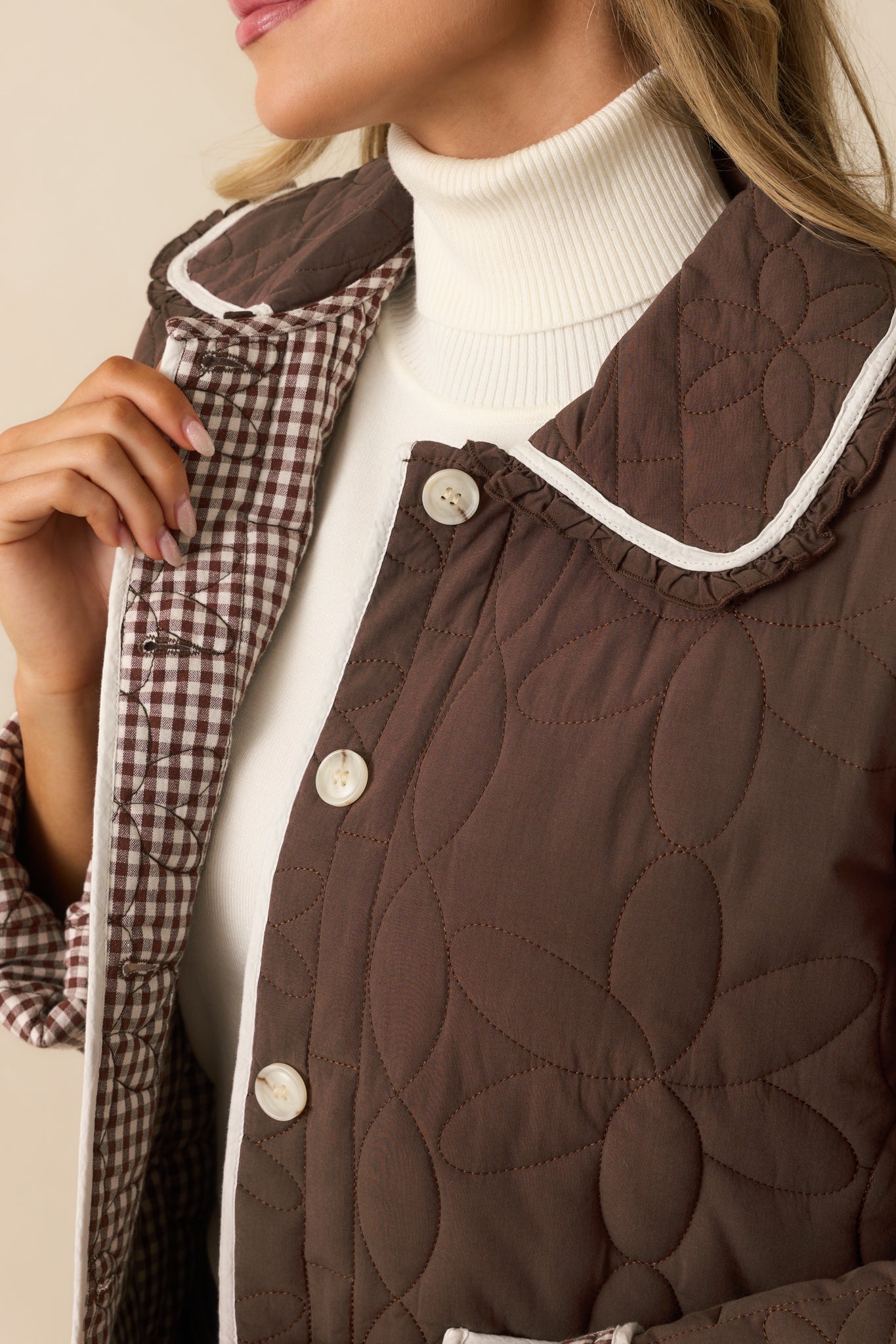 Up-close view showing the floral quilted stitching, ivory trim, and gingham lining of the brown jacket.