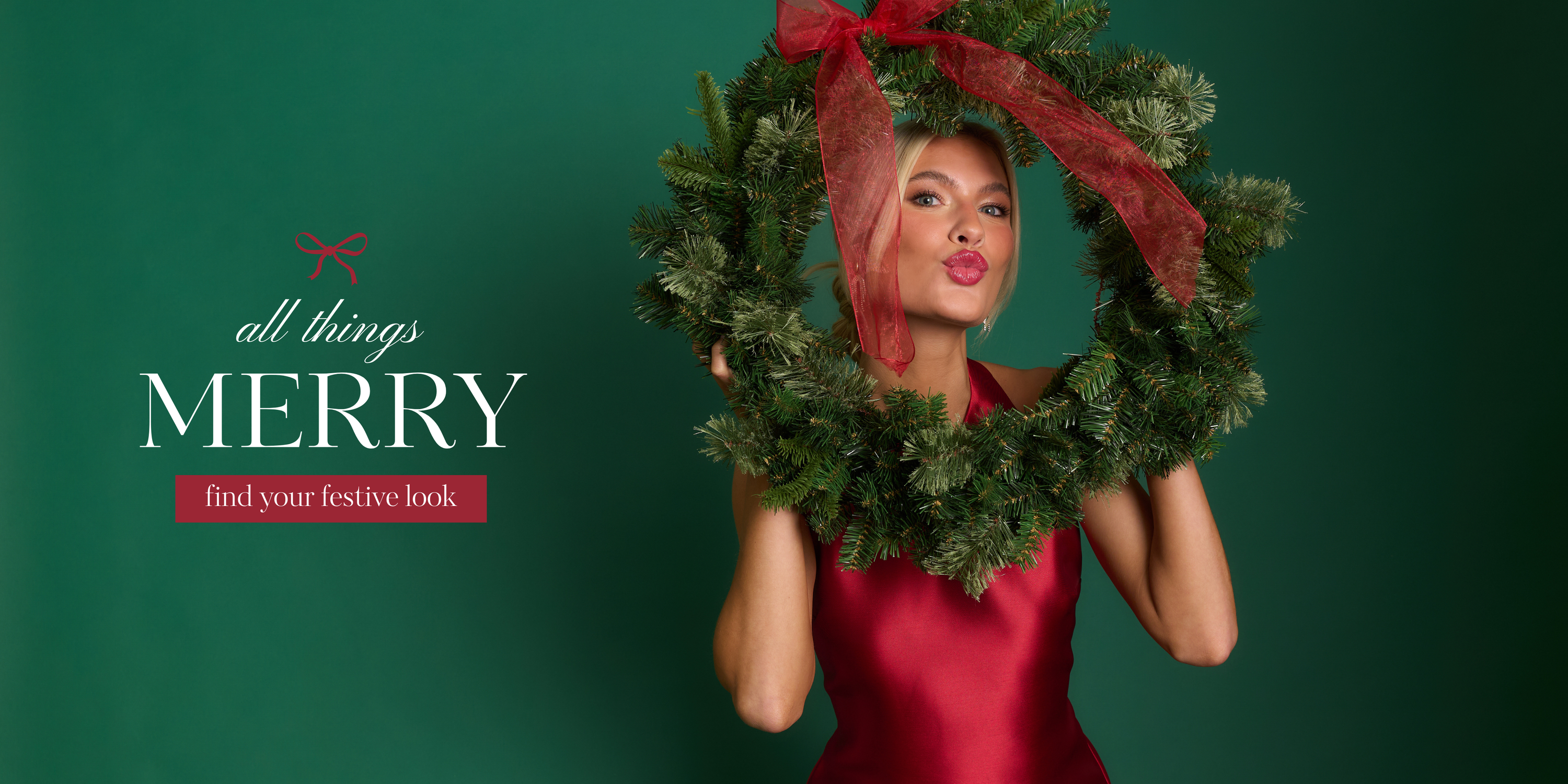 A model wearing a sleeveless red satin dress holds a large holiday wreath with a red bow, posing against a green backdrop. This dress is featured in our holiday shop.