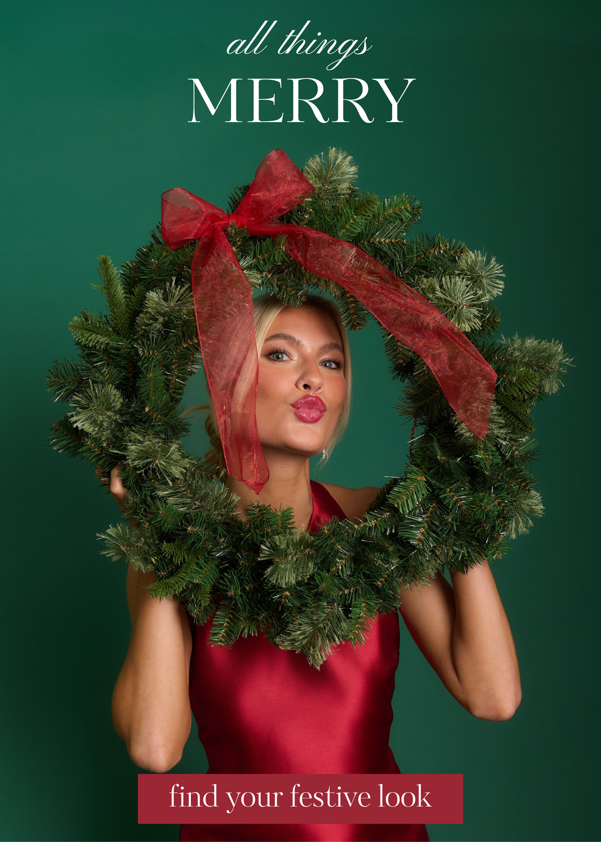 A model wearing a sleeveless red satin dress holds a large holiday wreath with a red bow, posing against a green backdrop. This dress is featured in our holiday shop.