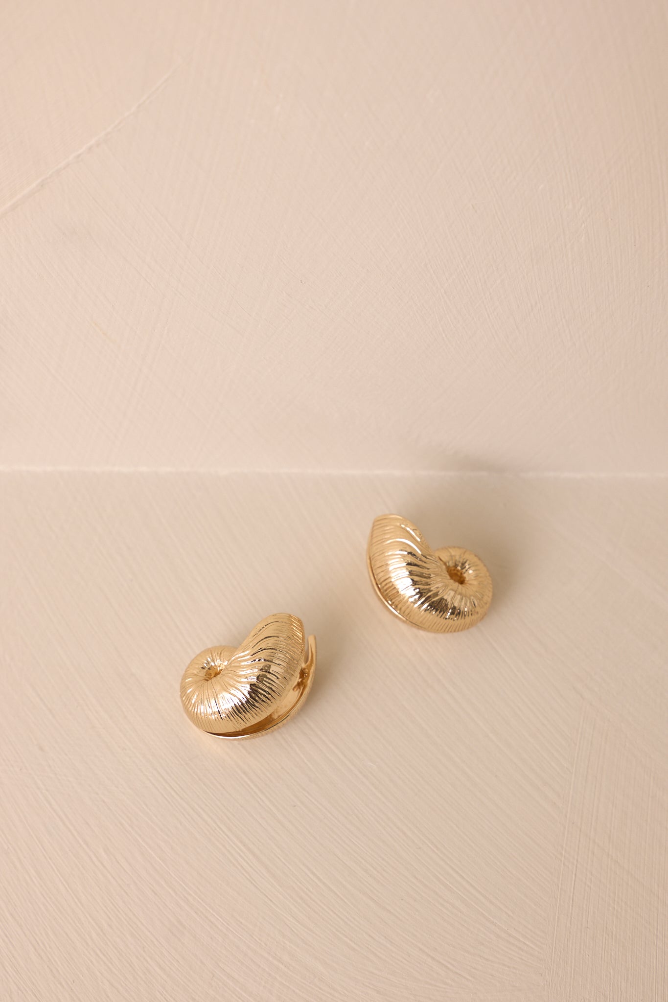 An angled view of the earrings on a beige background highlighting the shell-inspired curves and shine.