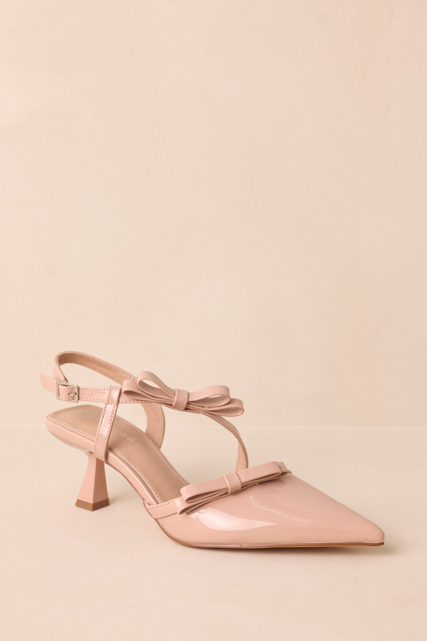 A slightly angled shot capturing the pointed toe, across-the-foot strap, ankle strap, and kitten heel against a beige background.