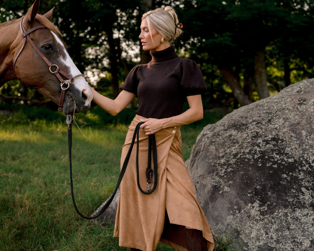 A woman stands outdoors in a grassy field next to a large rock, holding a horse lead rope while wearing a dark brown short-sleeve turtleneck top and a tan wrap-style skirt. A brown horse with a white blaze is visible on the left, and trees fill the background.