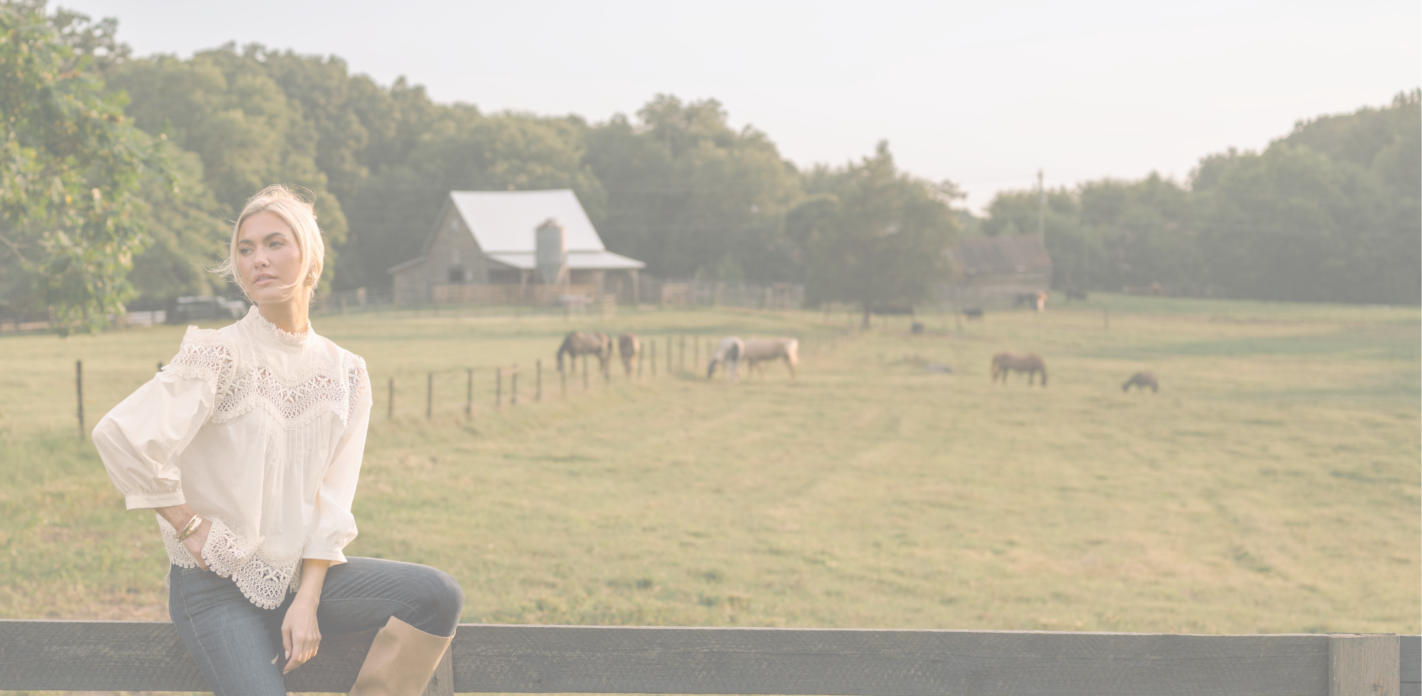 A woman wearing a white eyelet blouse and blue jeans sits on a fence in a rural setting with a barn, trees, and grazing horses in the background.