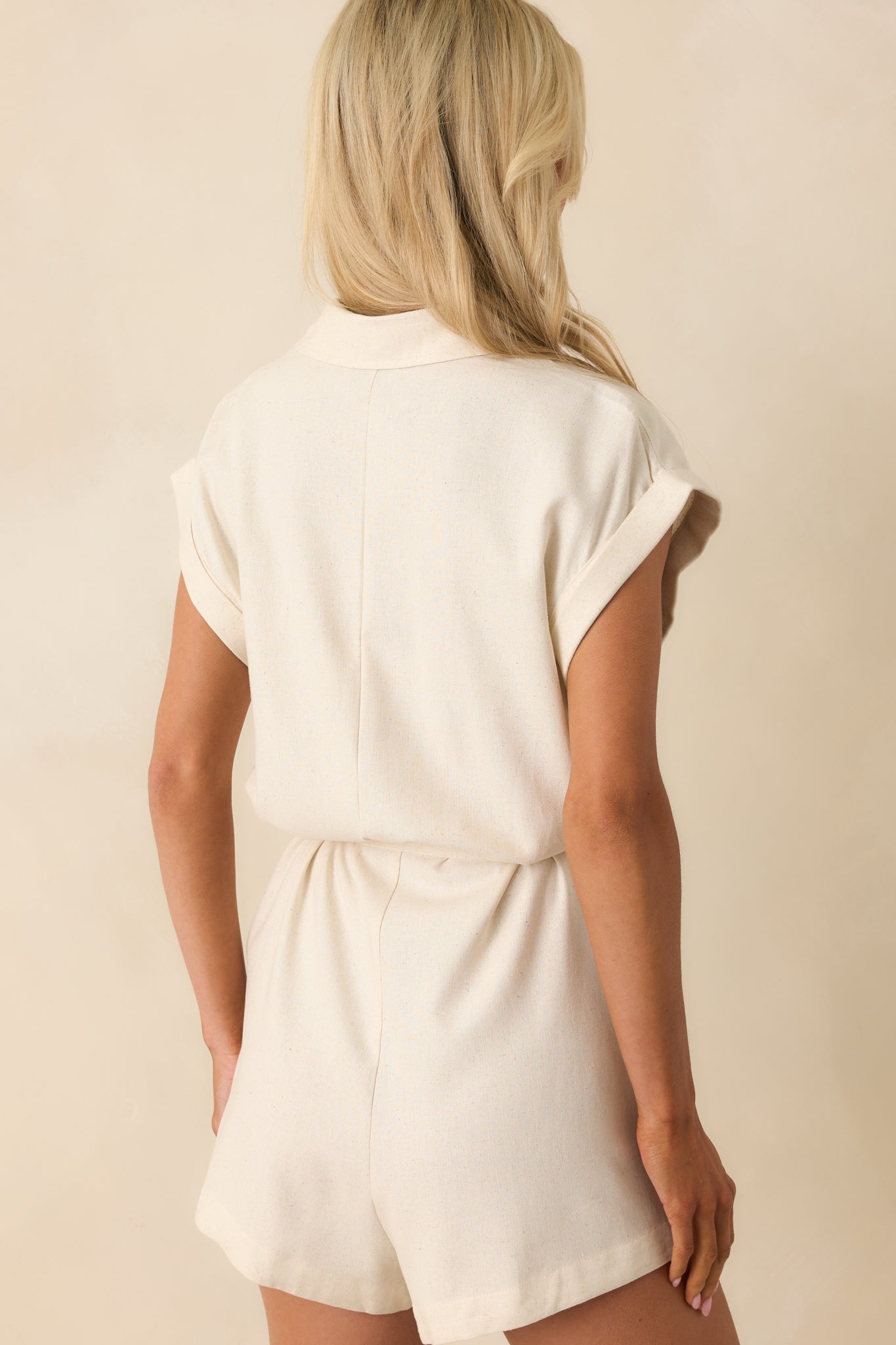 The natural ivory romper shown from the back, featuring a plain design with visible seams, relaxed fit, and the continuation of the cuffed short sleeves.