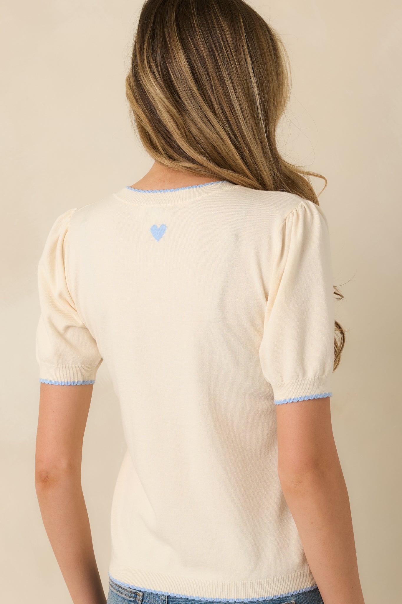 Back view of the ivory knit sweater highlighting the small blue heart detail centered at the back neckline and smooth knit finish.
