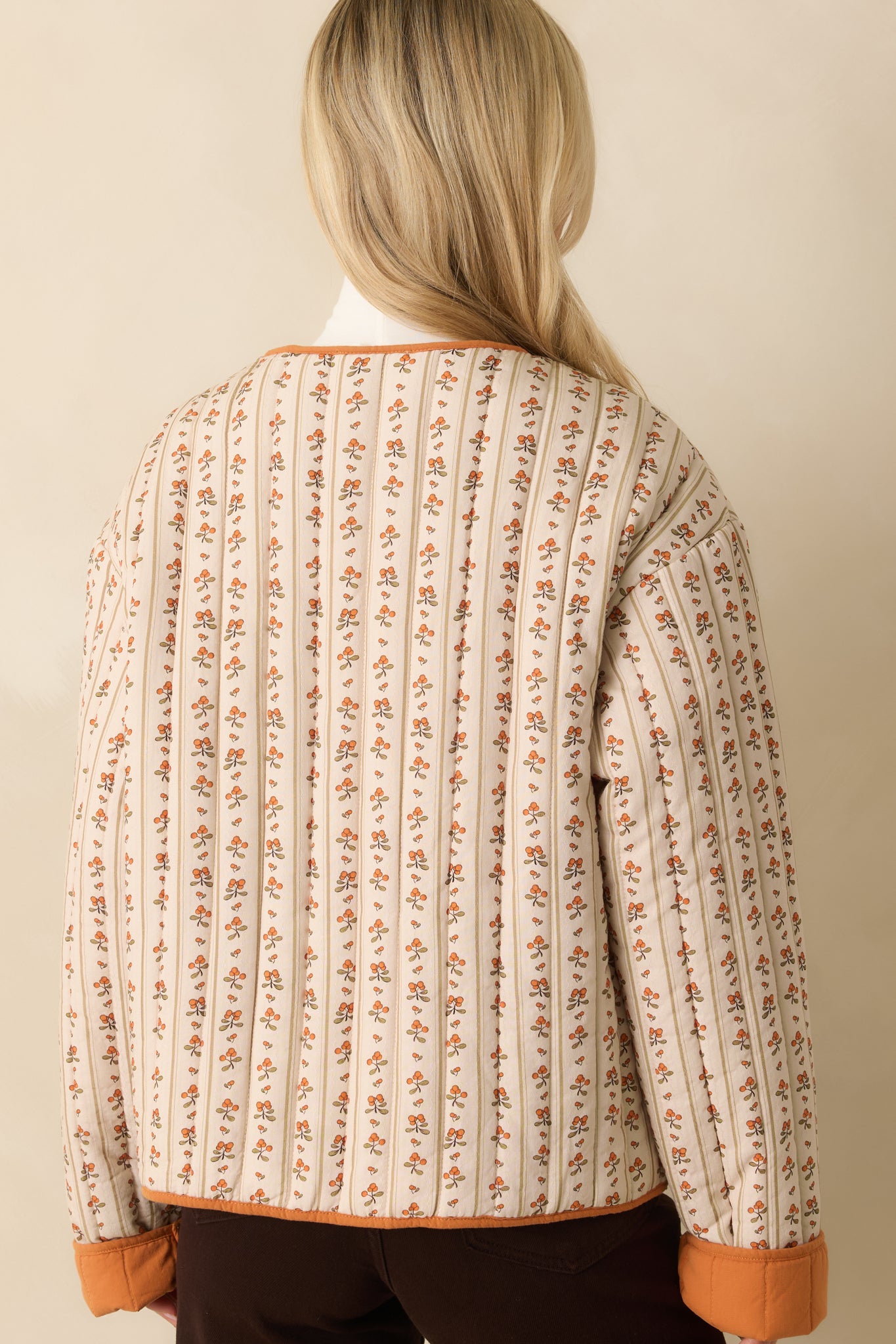 A back view of the ivory jacket featuring allover orange floral print, quilted texture, and contrast trim along edges.