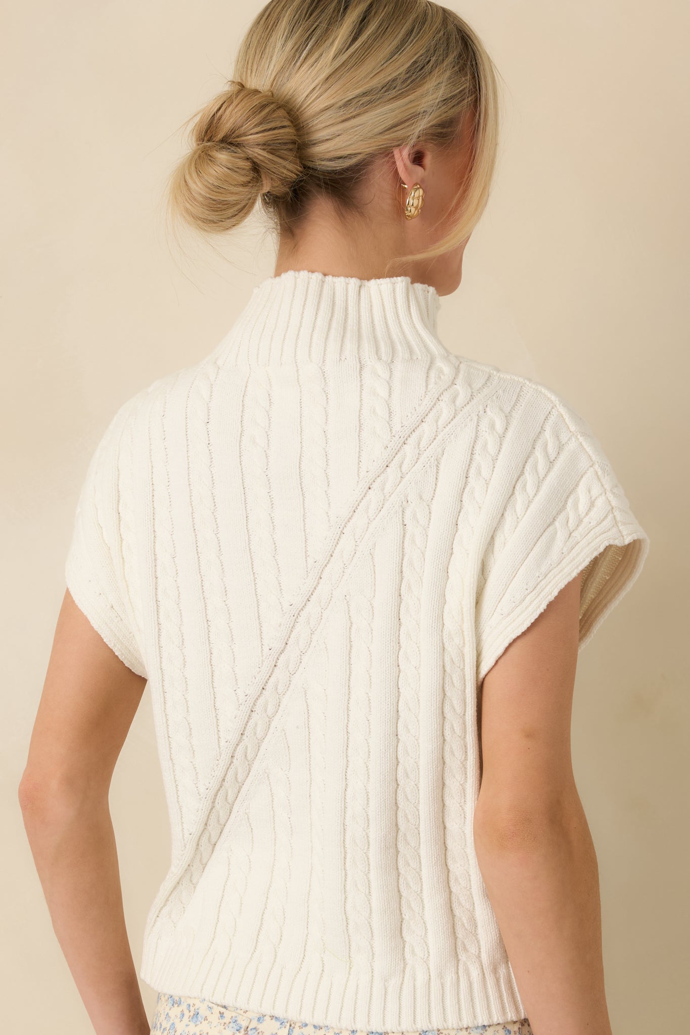 Back view of an ivory sleeveless mock neck sweater featuring a smooth knit finish and ribbed hemline.