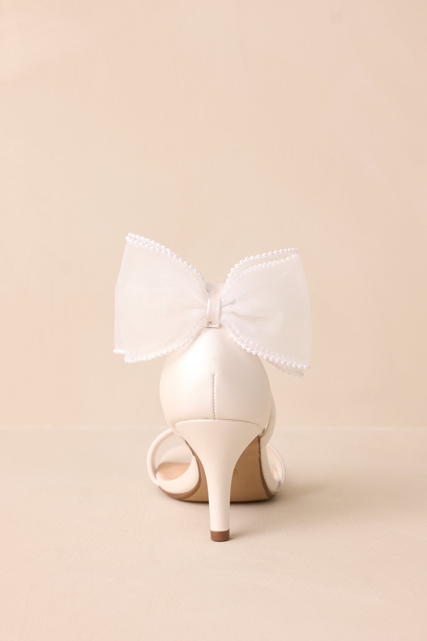 Back side view of the white heels, showcasing the ivory lace bow, the thin heel, and the secure ankle strap with silver buckle.