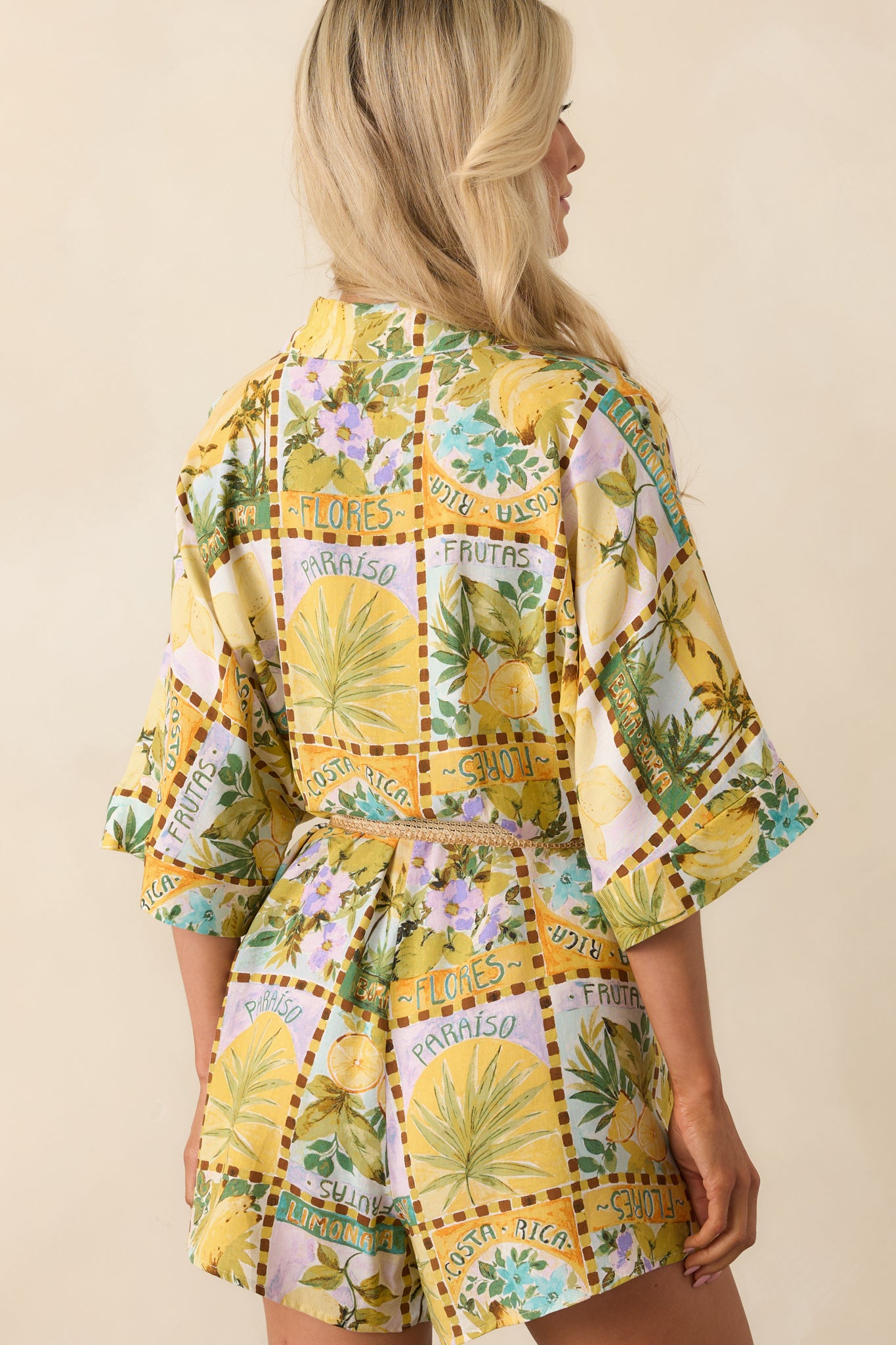 A view from behind displaying the allover fruit print, short sleeves, and belted waist of the yellow romper.