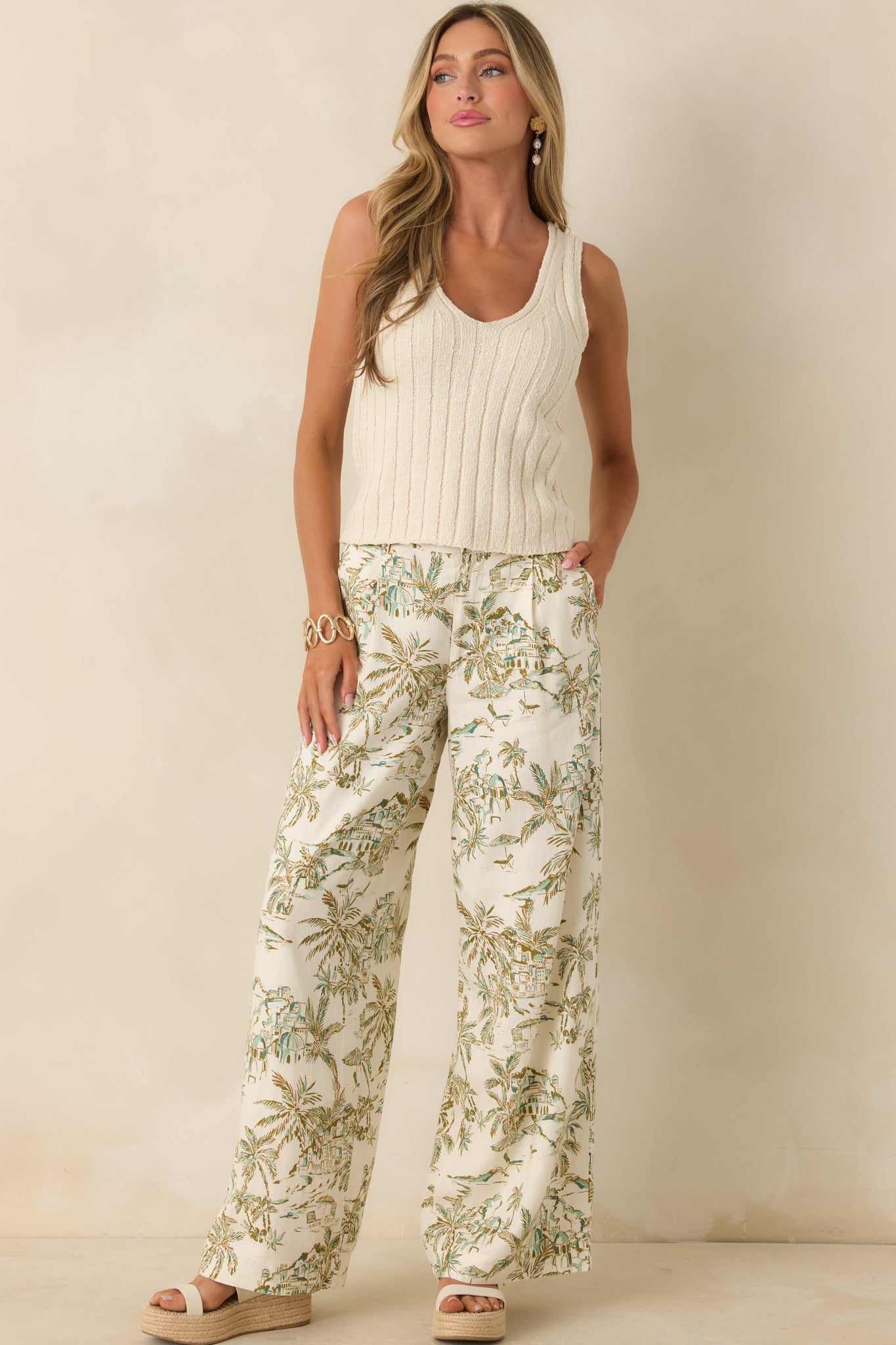 Light beige and green printed pants featuring a high-rise fit, wide leg silhouette, and full-length tropical scenic print in a washed linen blend.