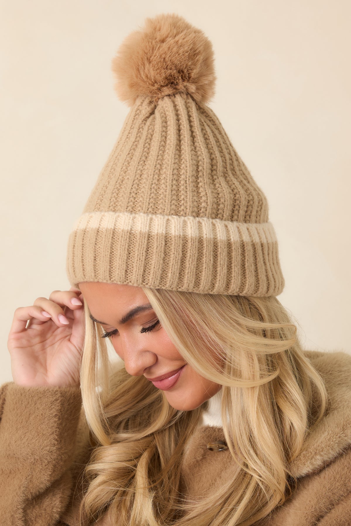 Neutral beige knit hat features a rounded slouchy crown, tall ribbed cuff, and fluffy pom in a coordinating tan shade.