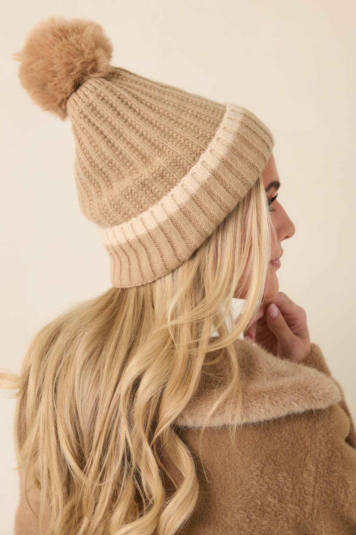 Beige knit hat displays a relaxed crown with pronounced vertical ribbing and a thick fold-over cuff beneath a fluffy tan pom.