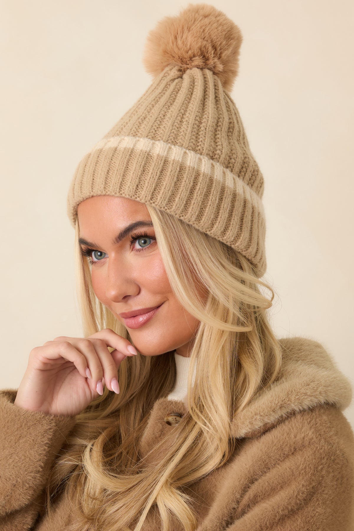 Beige knit beanie features vertical ribbing, a wide fold-over cuff, fluffy tan pom, and a relaxed slouchy crown.
