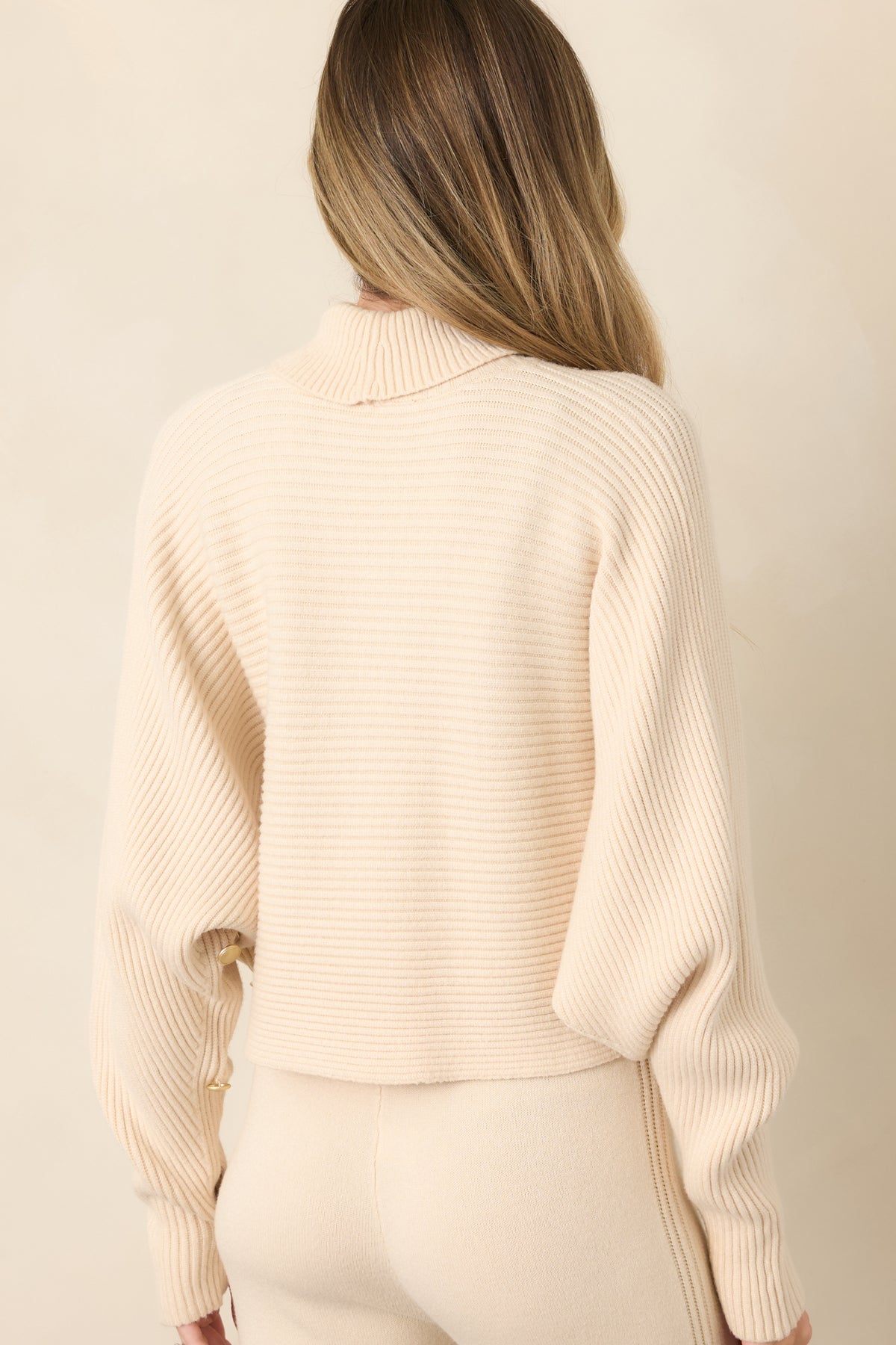 Rear view of the beige ribbed sweater highlighting the relaxed drape, wide batwing sleeves, high neckline, and cropped hemline.