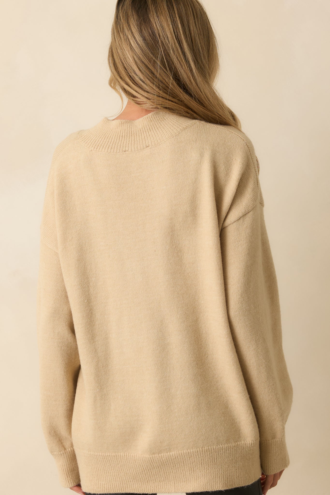 A back view of the beige sweater displaying plain knit fabric, oversized fit, and long sleeves with ribbed cuffs.