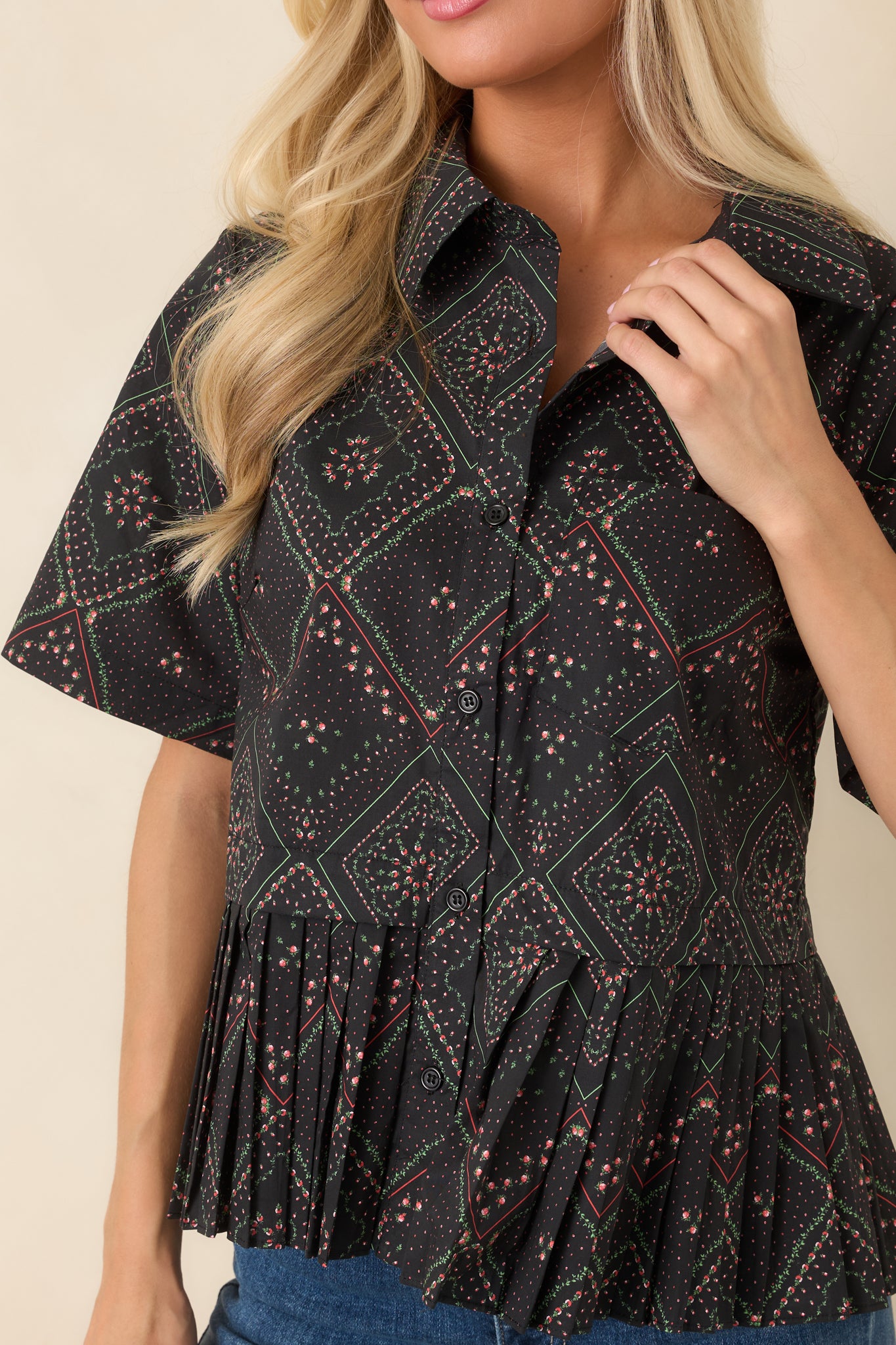 Close-up of the black cotton fabric featuring coral and green floral motifs within a scarf-inspired geometric print, complemented by the collared neckline, functional buttons, and crisp pleated construction.