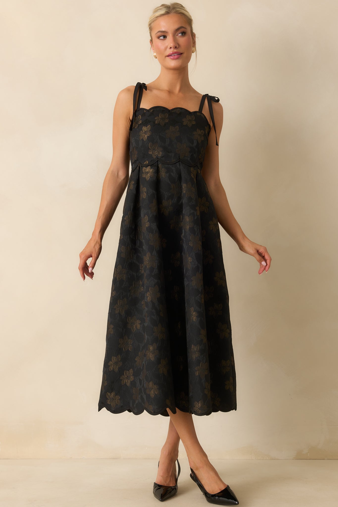 Black midi dress with gold floral jacquard pattern, scalloped hem, and self-tie shoulder straps, showing the fitted bodice and front pockets.