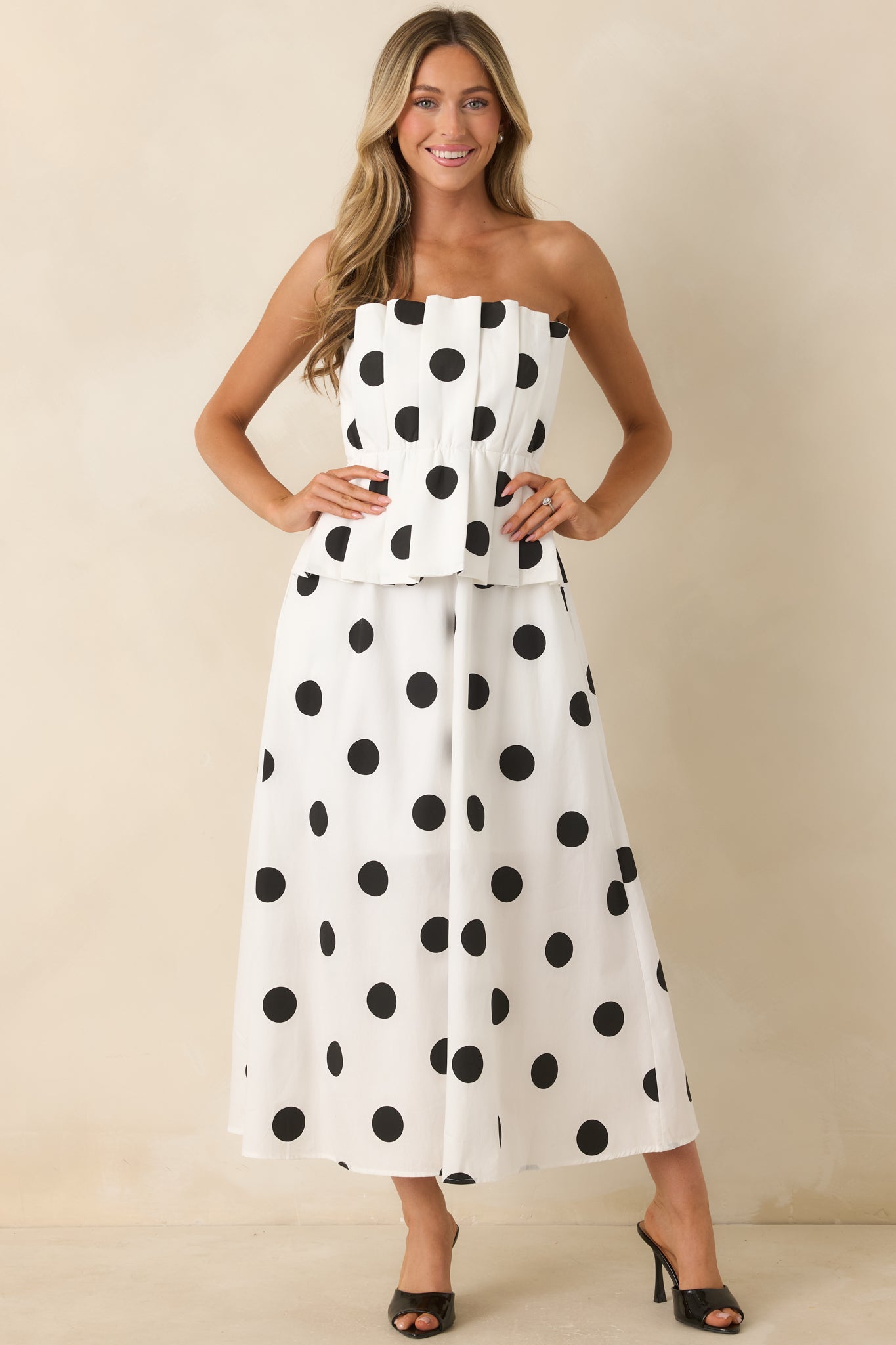 Black polka dot midi dress with a strapless neckline, sculptural pleated waist overlay, hidden side pockets, and a graceful A-line silhouette.
