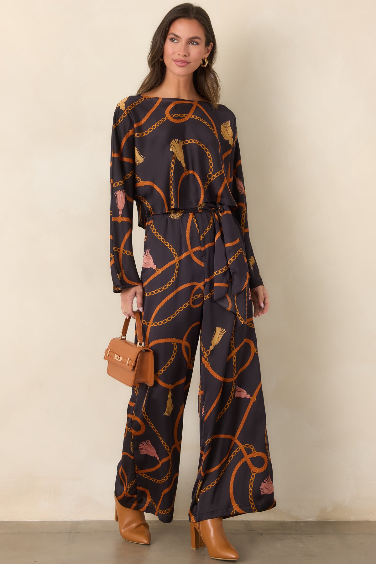 Black printed satin-feel pants showcasing warm rust and gold motifs, a flowing wide-leg cut, and a matching tie belt.