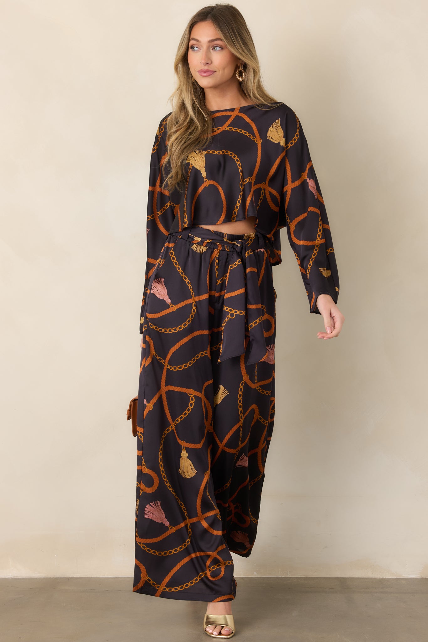 Black satin-feel pants featuring a warm rust, gold, and blush rope, chain, and tassel print with a relaxed wide-leg silhouette.