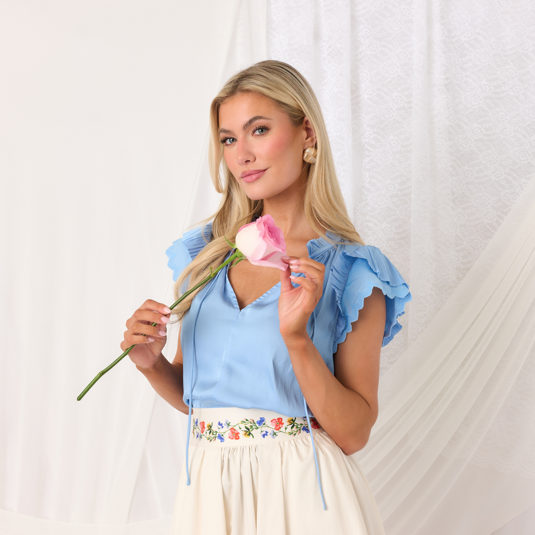Woman holding a pink flower against a white background, wearing a blue flutter sleeve, silk like blouse. 