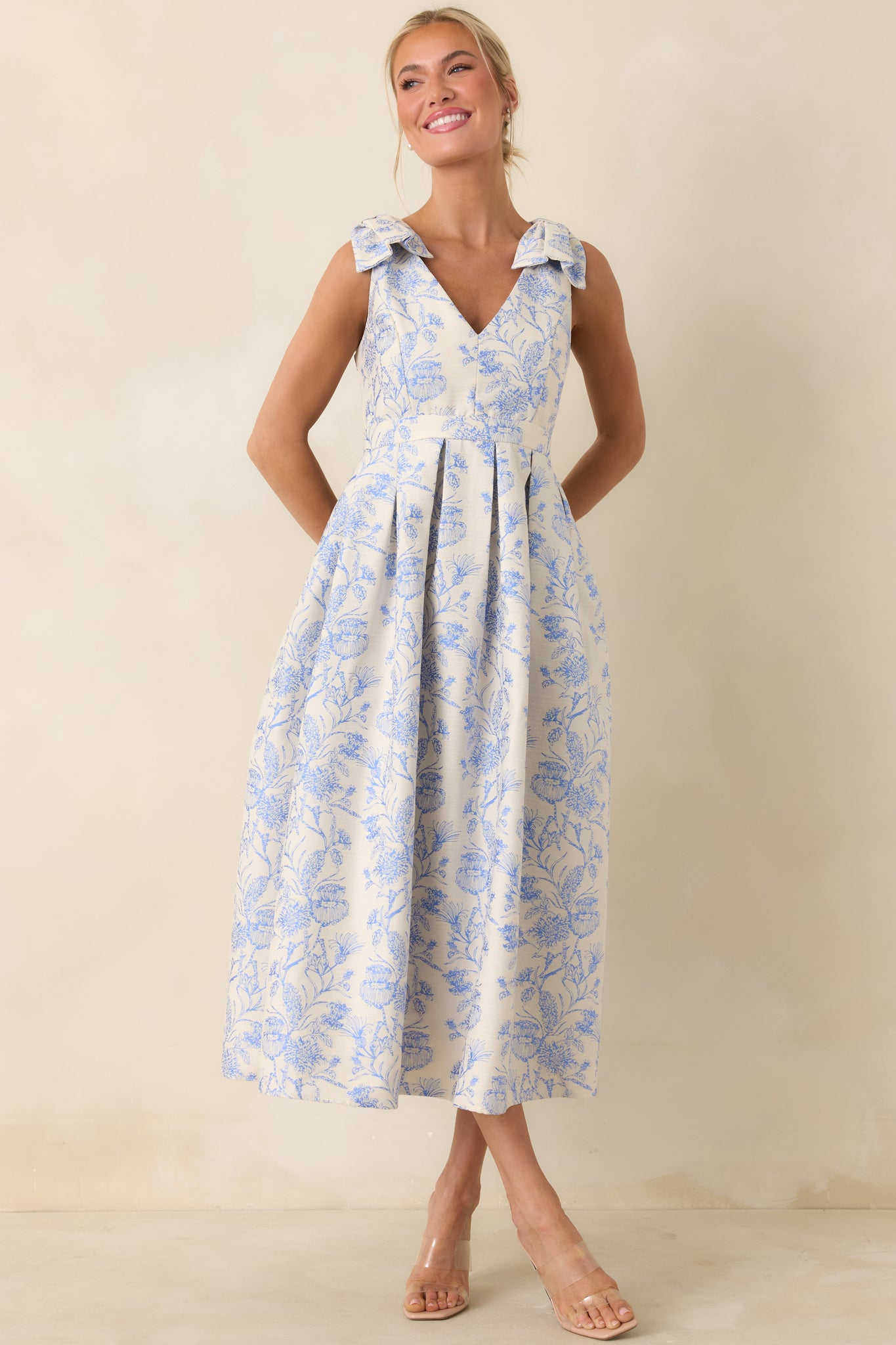 Front view of a blue floral midi dress with bow-tie shoulder straps, V-neckline, and pleated skirt.