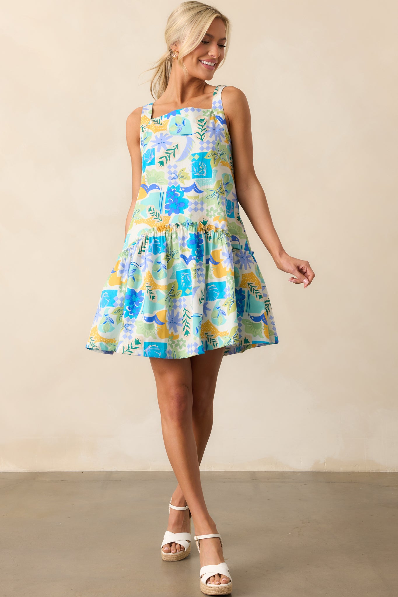 A vibrant blue mini dress with a multicolor tropical print, featuring a high square neckline, ruffle accents, and a breezy drop waist design.