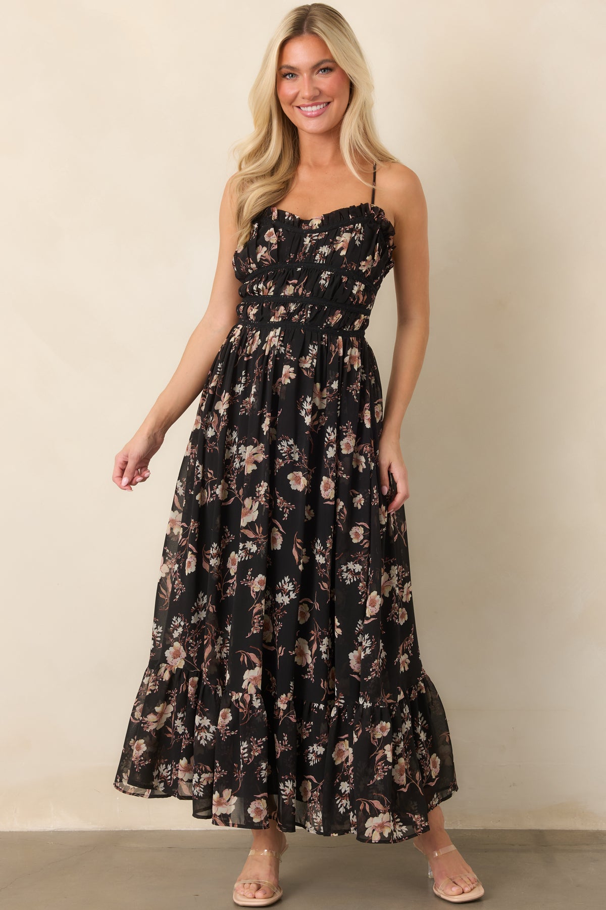 Blush and ivory blooms pattern the sheer black dress, highlighting its gathered bodice, slender straps, and flowing ruffled hem.