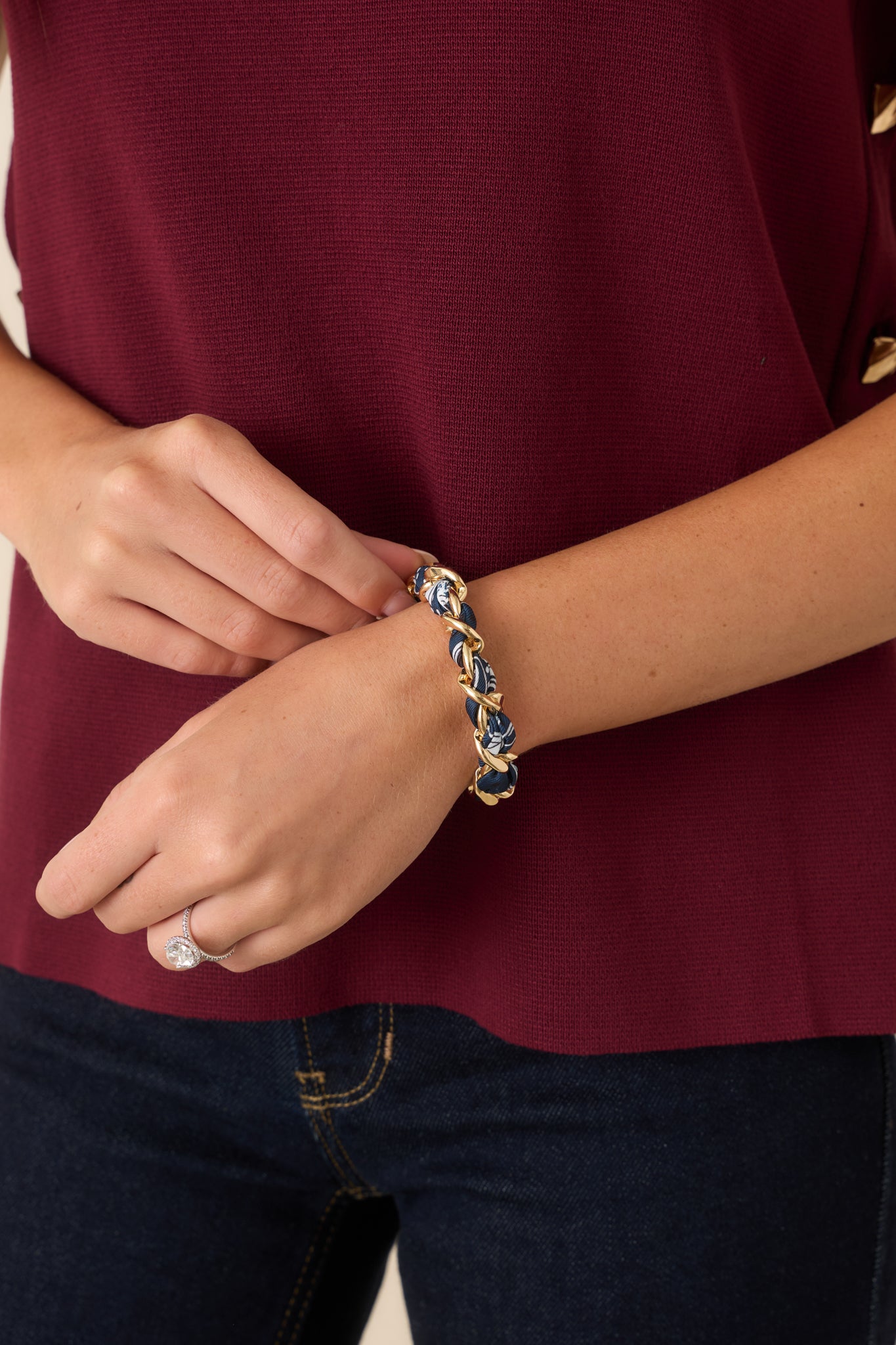A close-up of a person wearing a braided blue scarf and gold link bracelet, paired with a burgundy top and dark blue jeans.