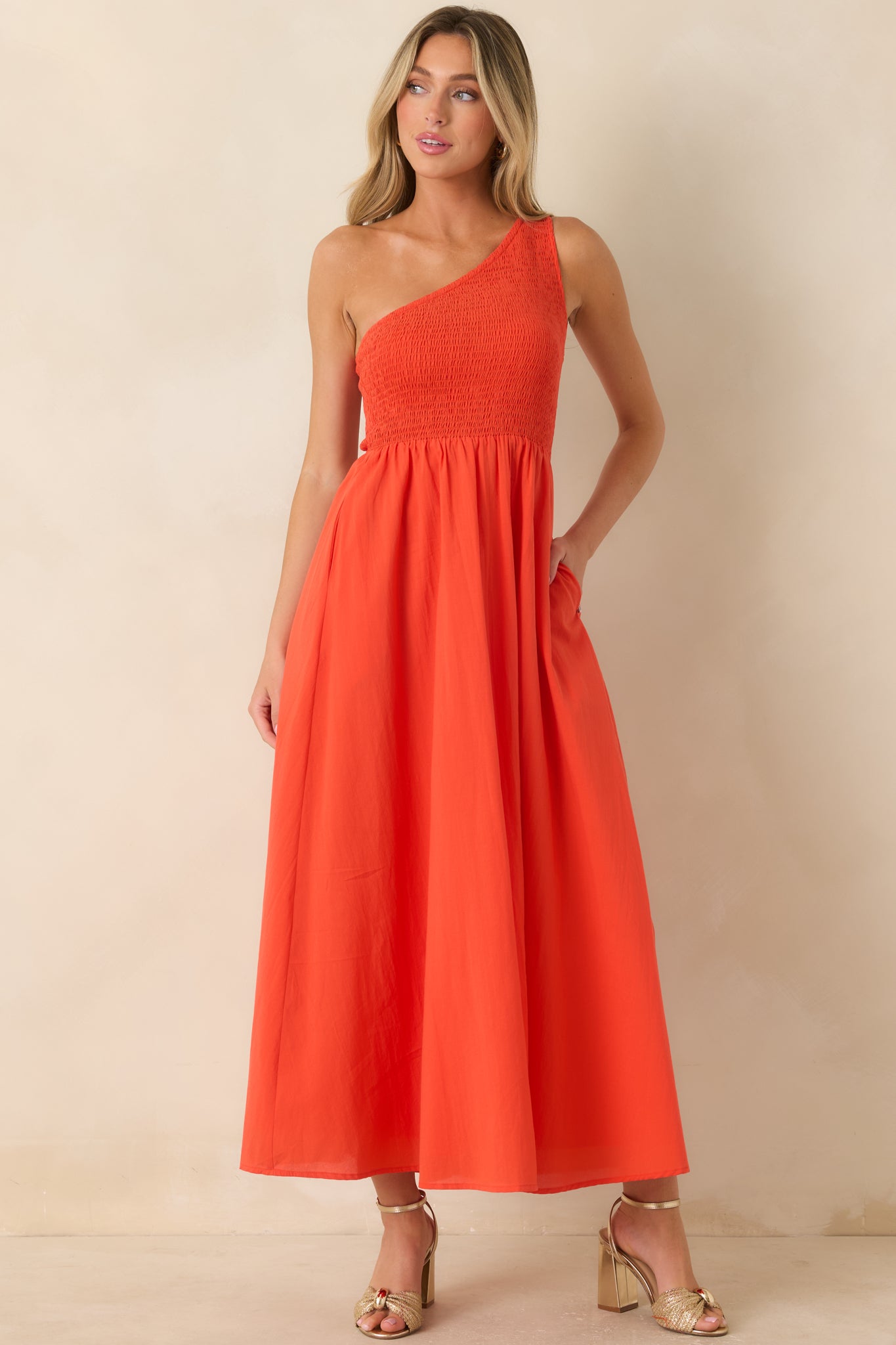 Bright coral-orange maxi dress featuring a textured smocked bodice, asymmetrical neckline, hidden side pockets, and a graceful full-length skirt.