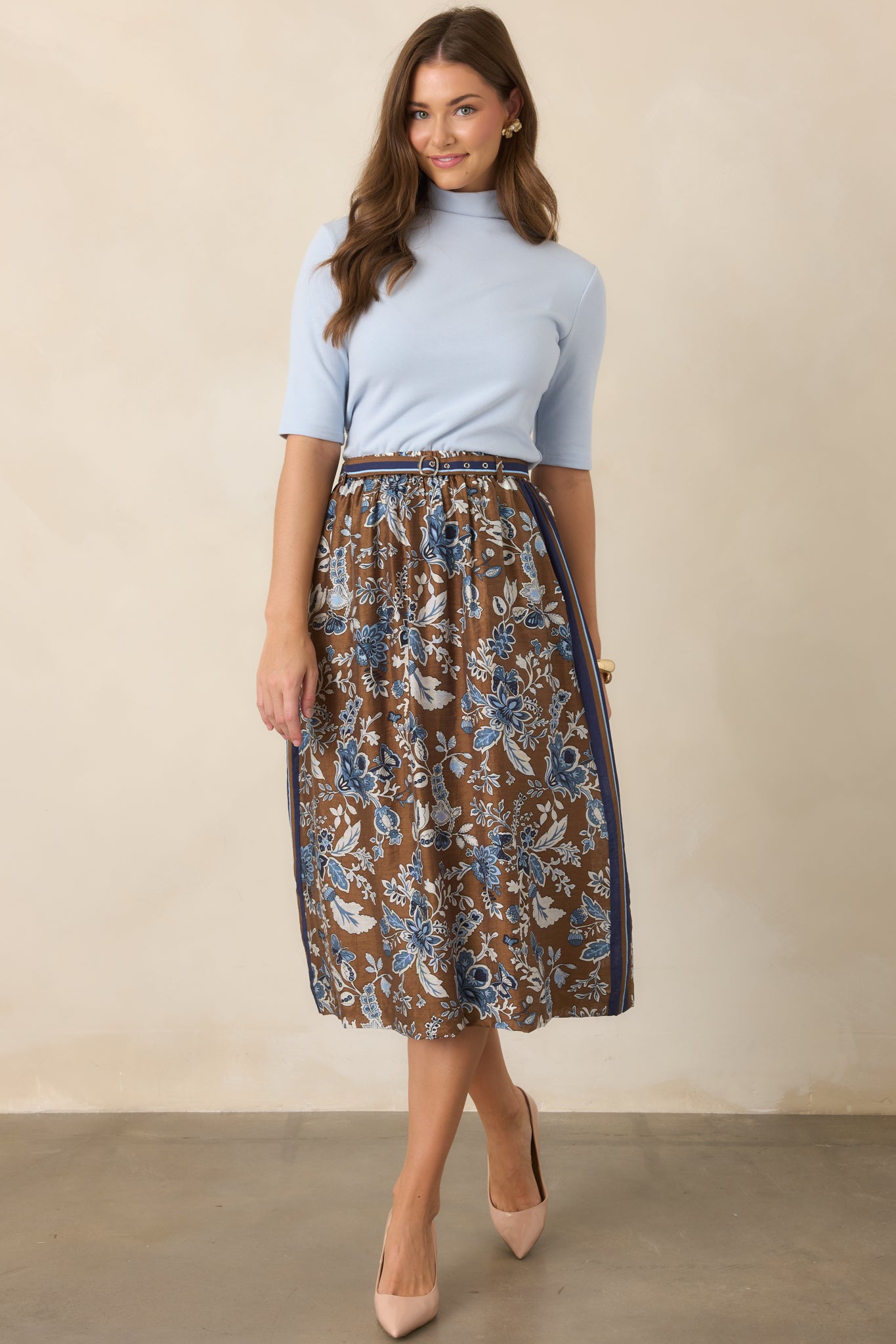 Rich brown A-line midi skirt covered in blue floral motifs, finished with striped trim along the waist and sides, a coordinating double-buckle belt, and functional pockets.