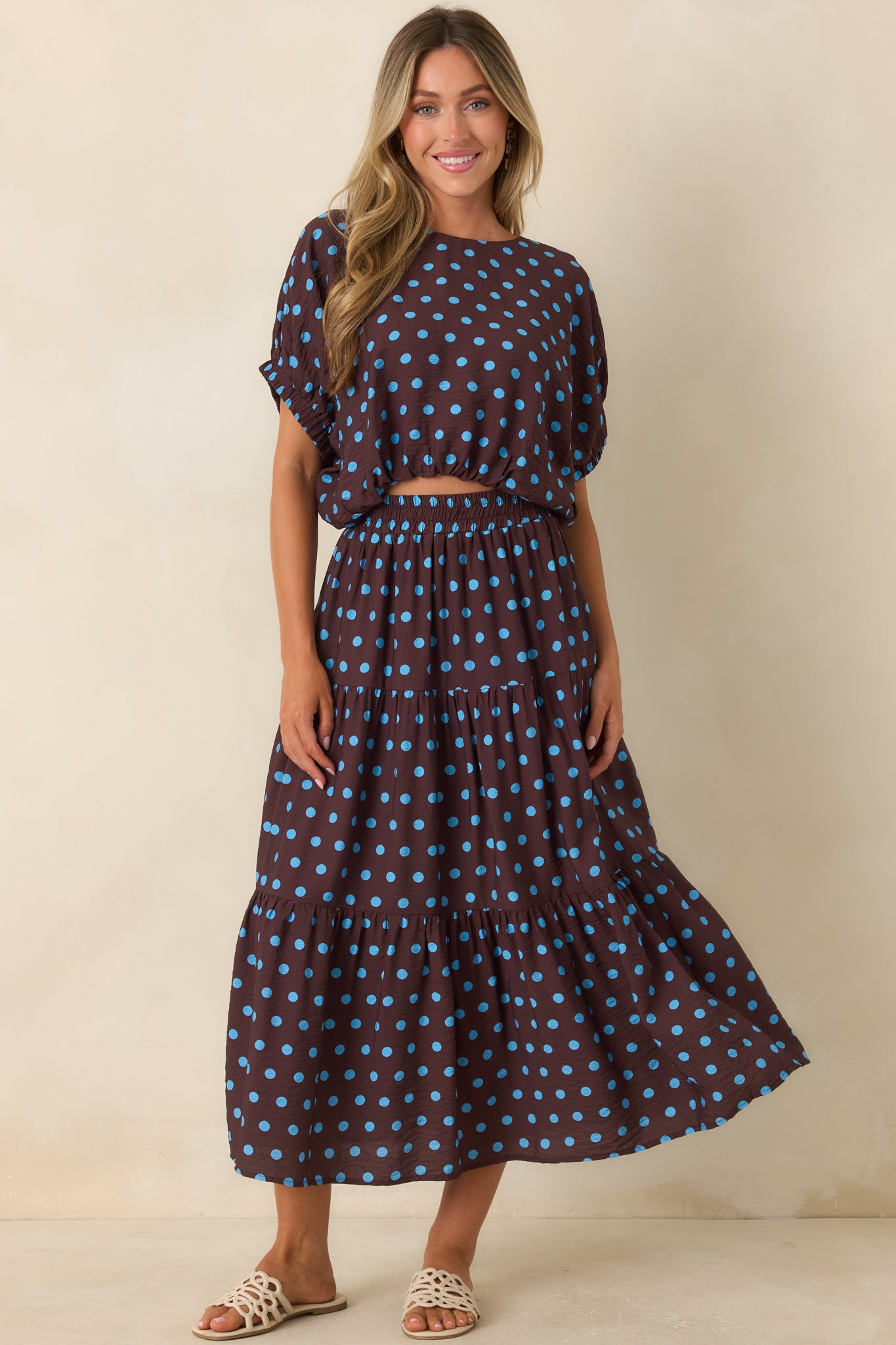 Brown blouse featuring vibrant blue polka dots, a round neckline, draped short sleeves, and a relaxed blouson silhouette.