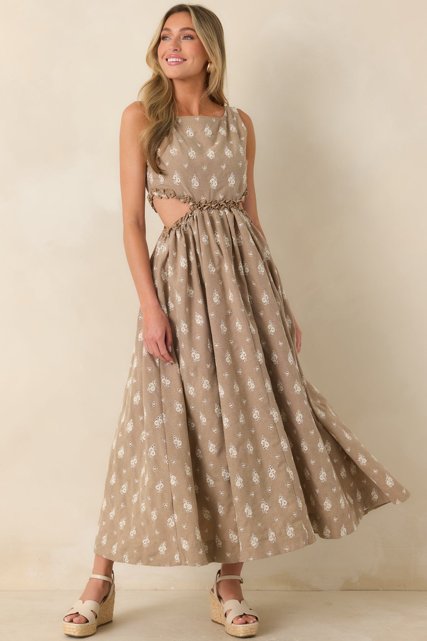 Full-length view of a sleeveless maxi dress featuring a brown and cream floral eyelet print, boat neckline, gathered waist, and flowing skirt with side cut-outs and ruffle trim.
