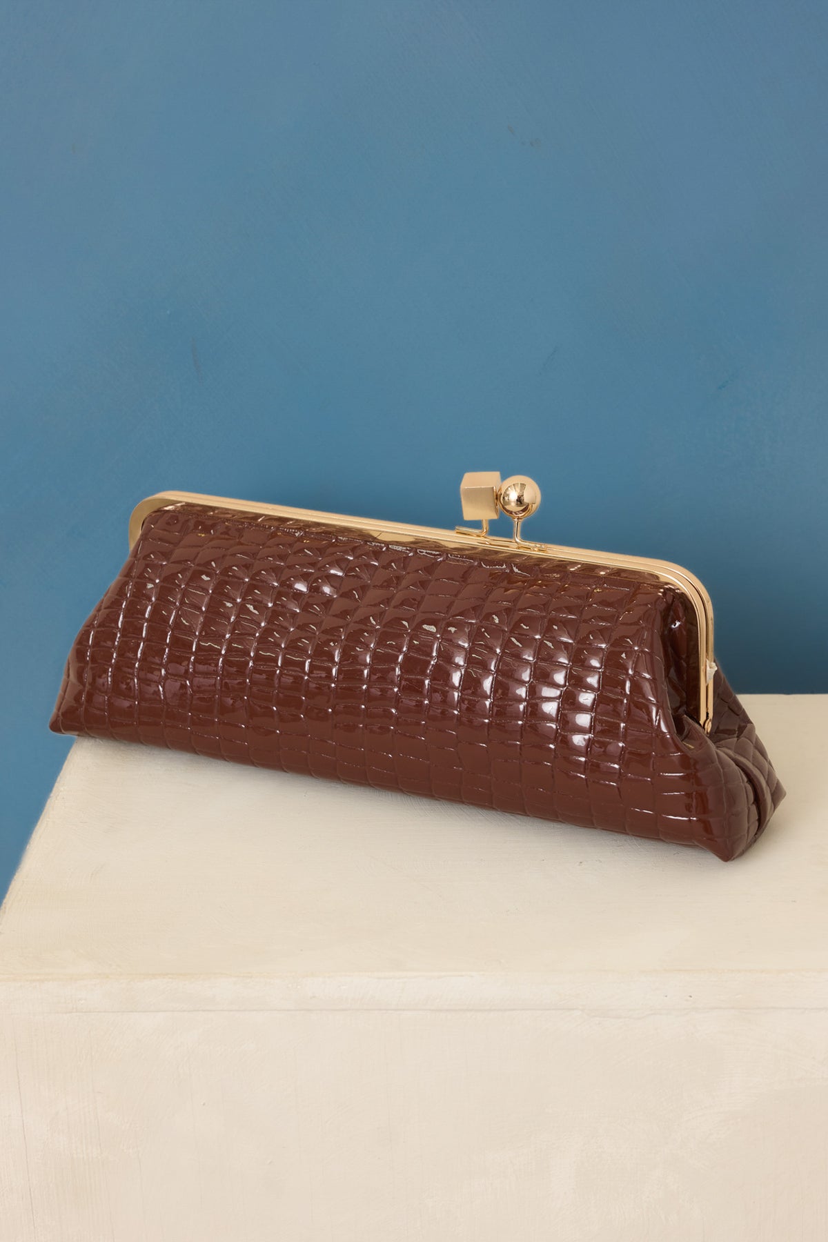 Brown clutch highlighting the raised croc-embossed texture, rectangular shape, and gleaming gold-tone kiss-lock closure on blue back drop.