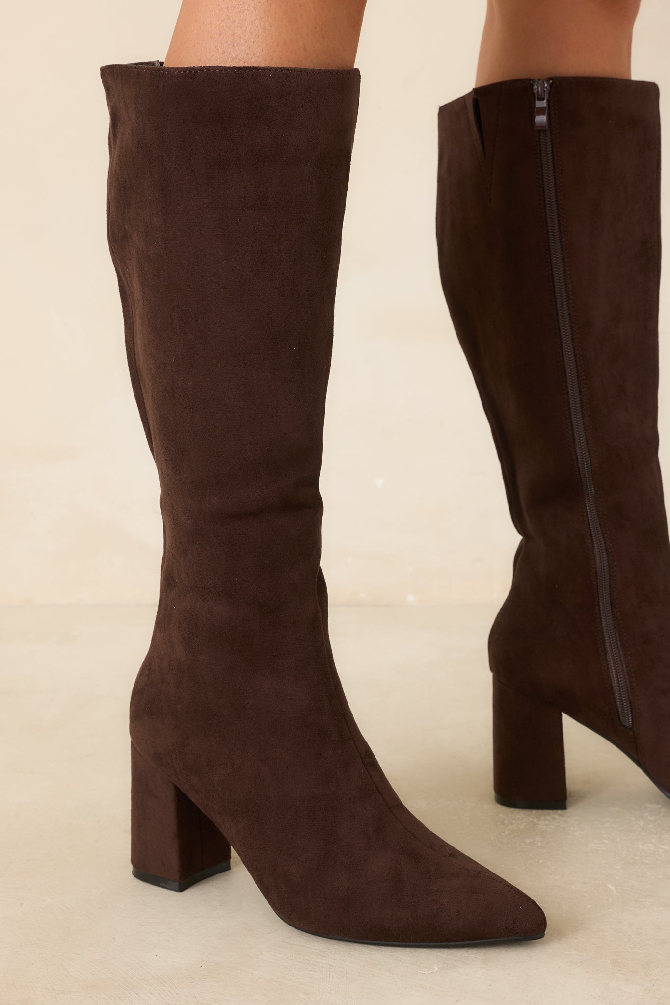 Deep brown faux suede tall boots featuring a pointed toe, fitted knee-high shaft, covered block heel, and smooth streamlined silhouette against a beige background.