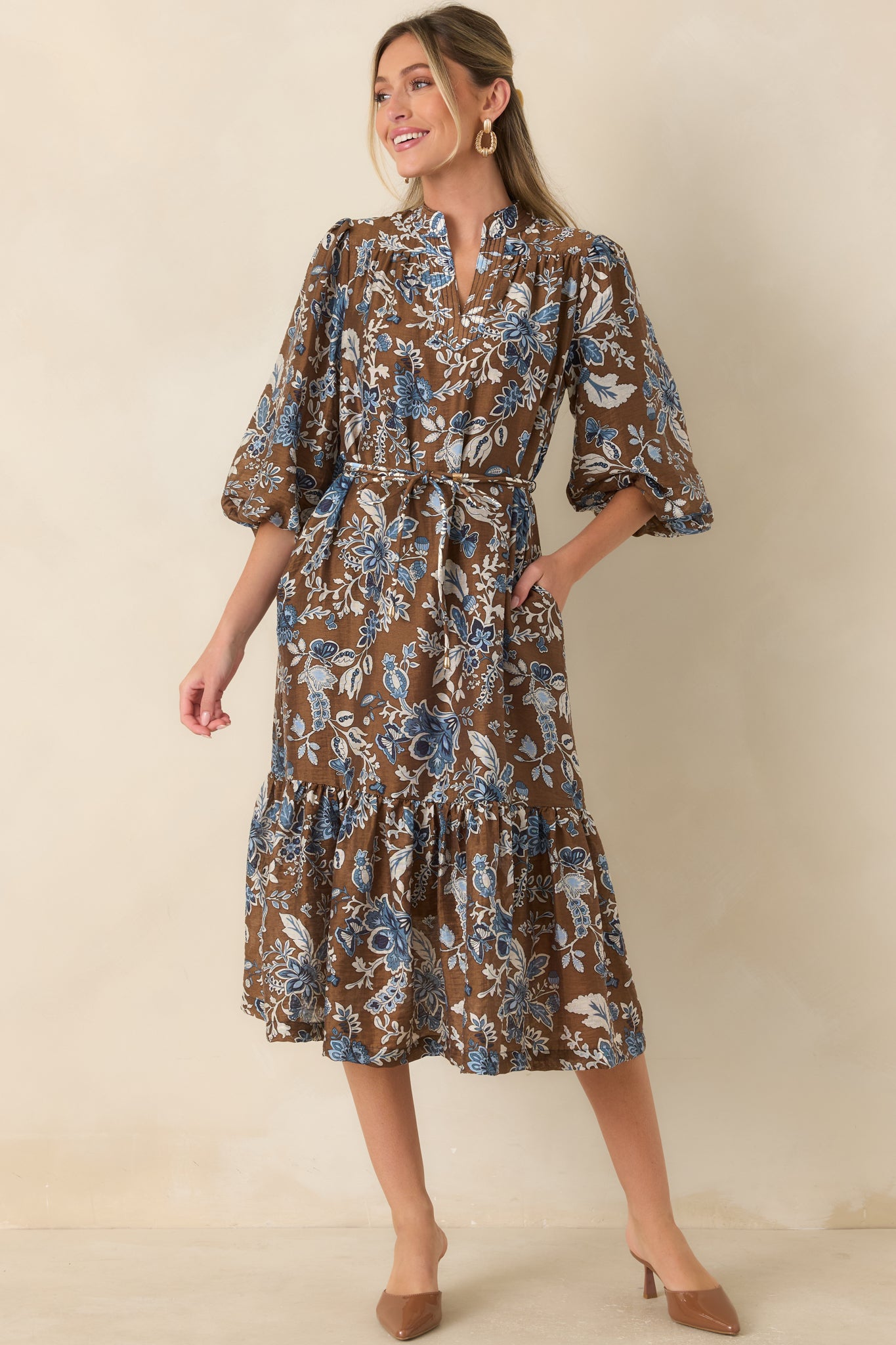 Floral midi dress featuring a warm brown background, blue floral print, split band collar, tie belt, and gathered ruffle hem.