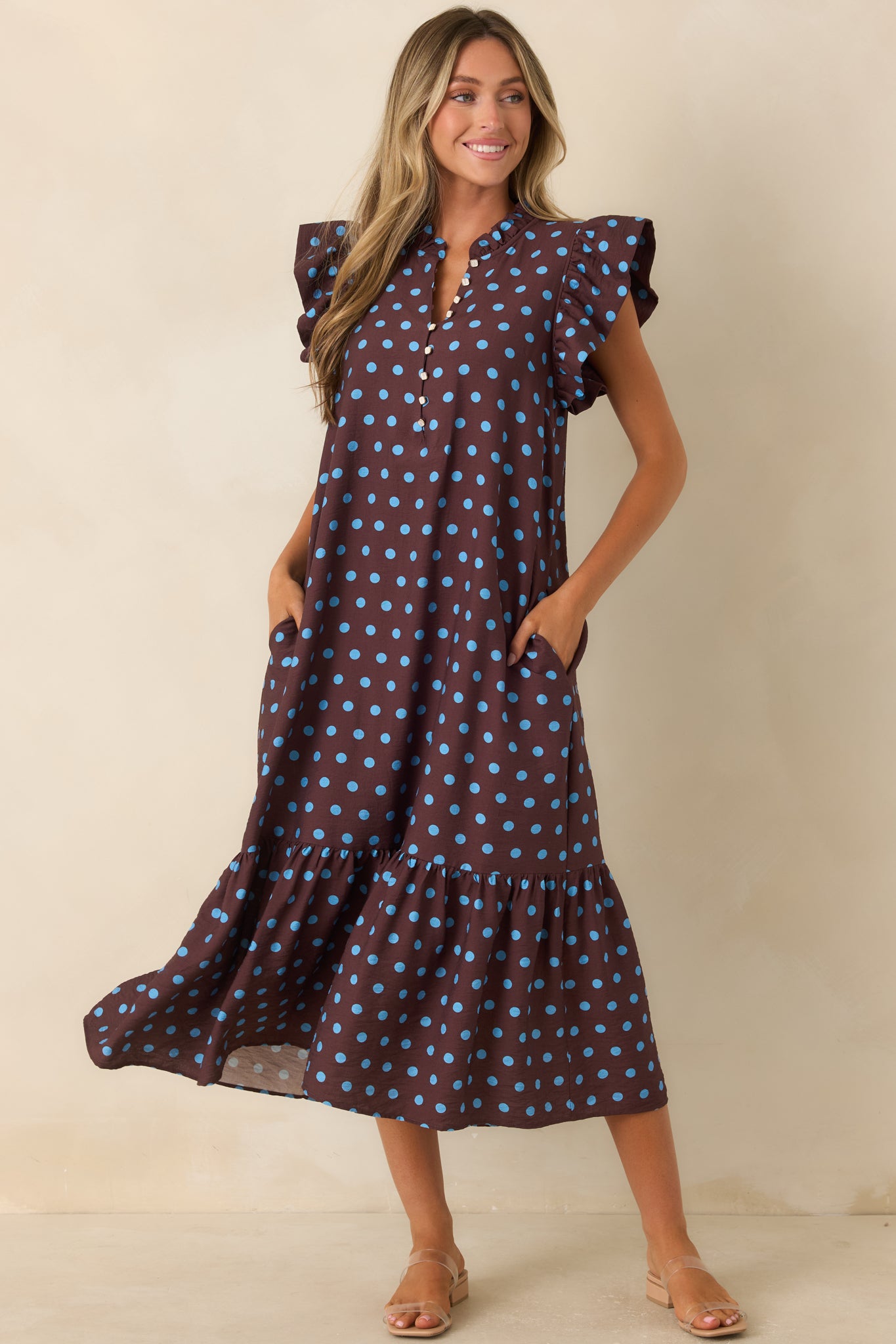 Brown midi dress featuring vibrant blue polka dots, flutter sleeves, pearl-inspired buttons, ruffle trim, and a tiered hem.