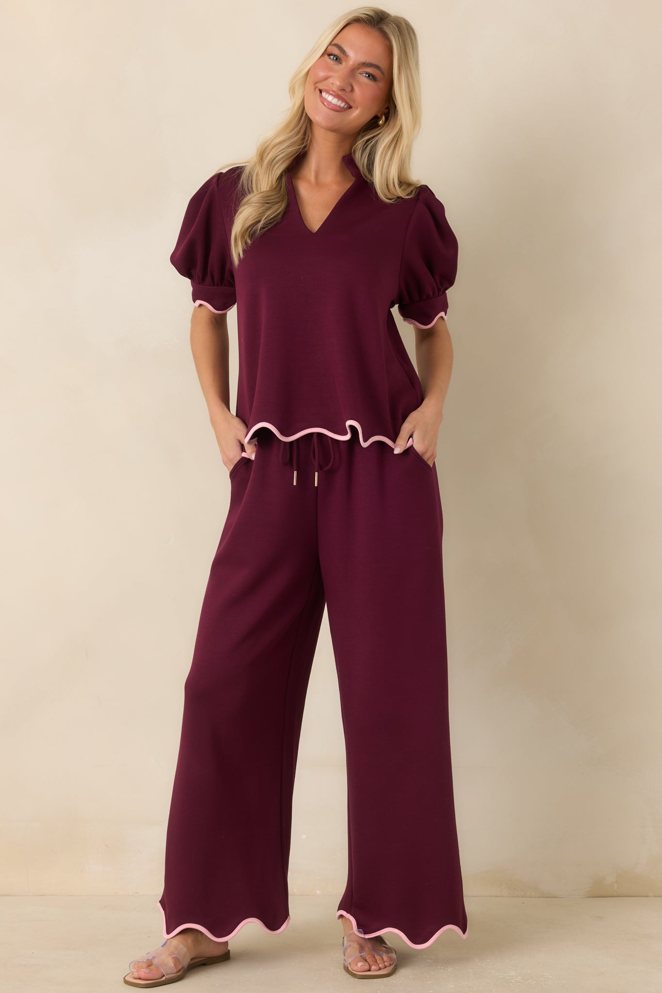 Burgundy knit pants featuring a relaxed wide leg silhouette, an adjustable drawstring waistband with gold-tone cord ends, and blush pink scalloped trim at the hem.