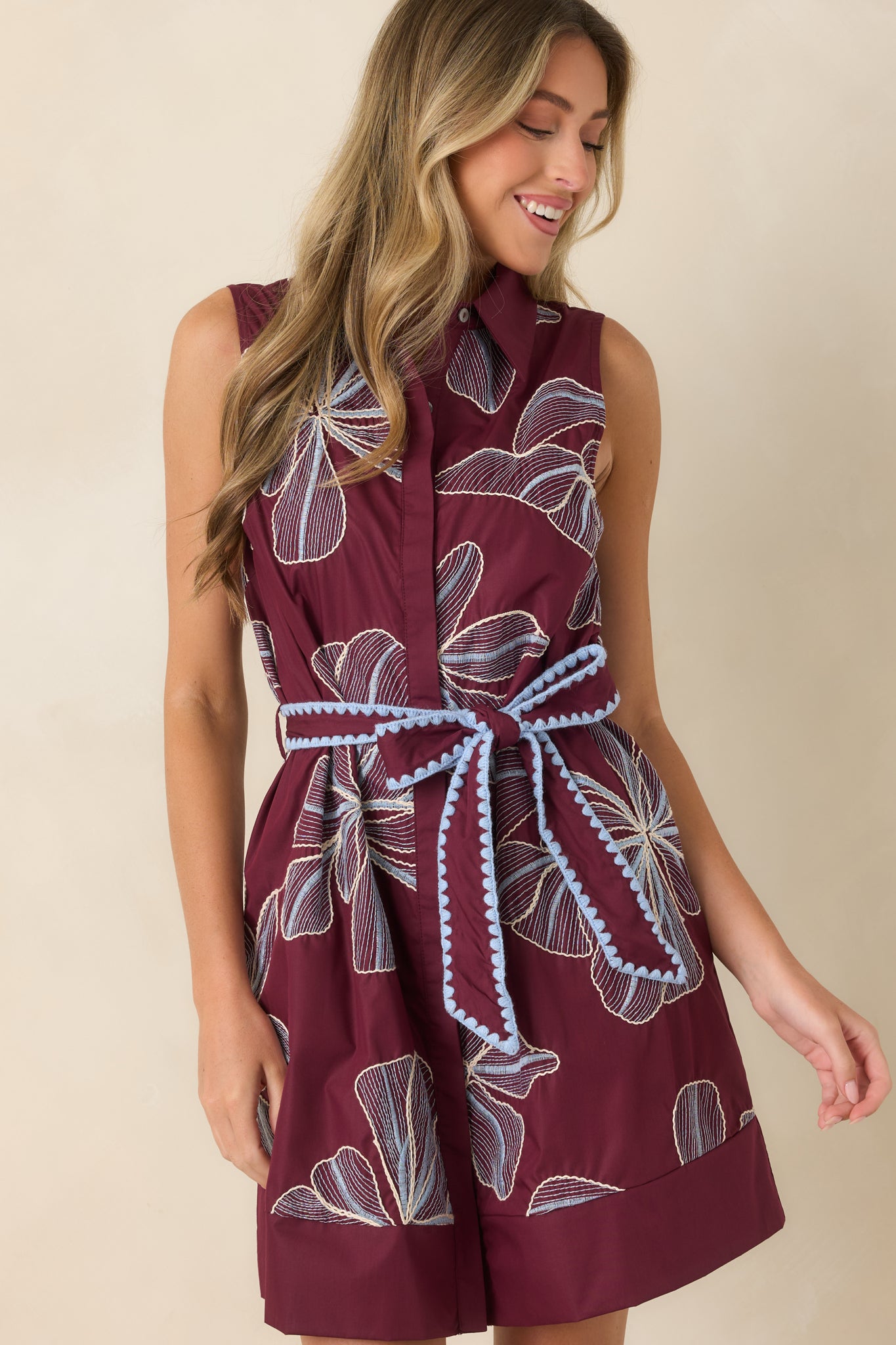 Detailed view of the burgundy mini dress featuring oversized embroidered florals and soft blue scalloped trim along the sash belt.