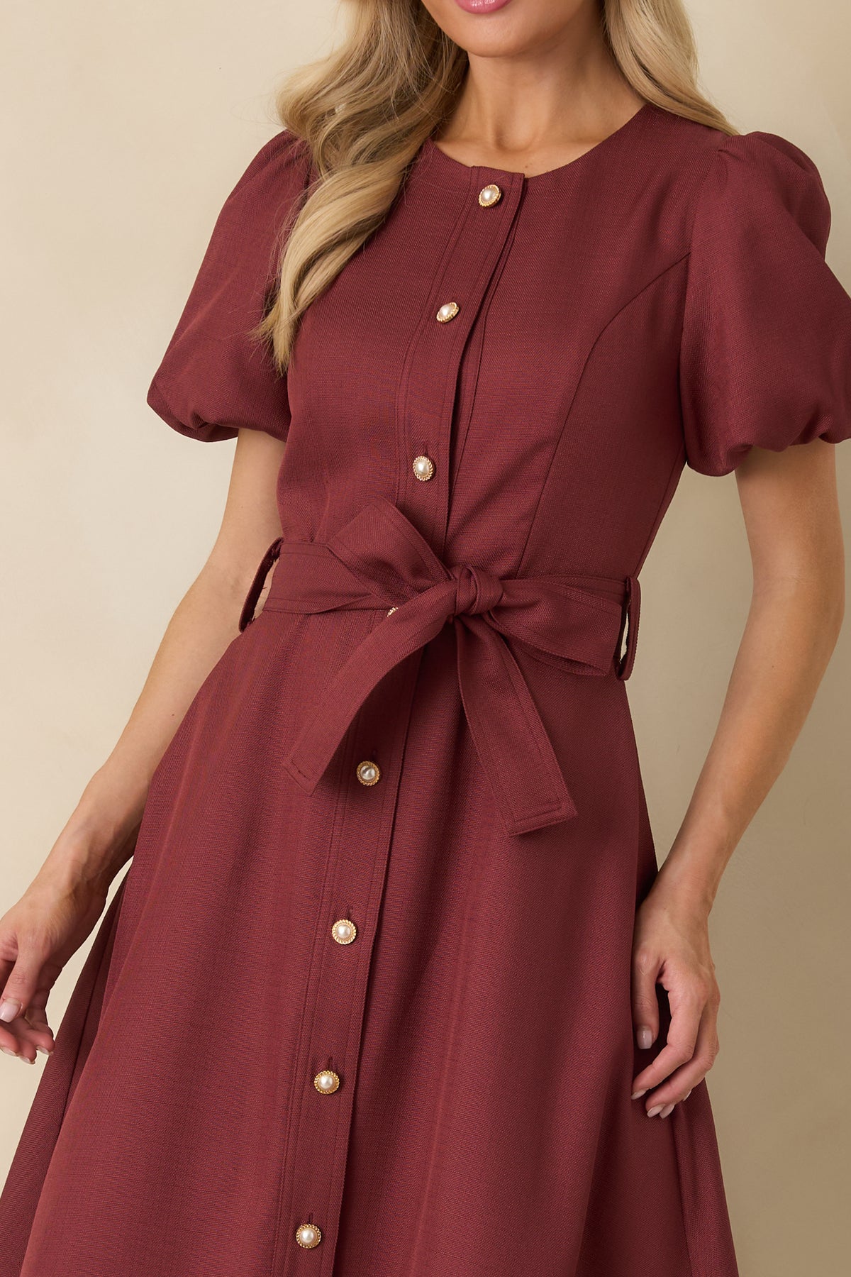 Close-up of burgundy woven dress featuring a round neckline, pearl and gold buttons, puff sleeves, and self-tie belt.