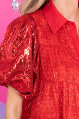 Close up view of this dress that features a collared neckline, functional red buttons down the front, sequin puff sleeves, and sparkly tinsel throughout.