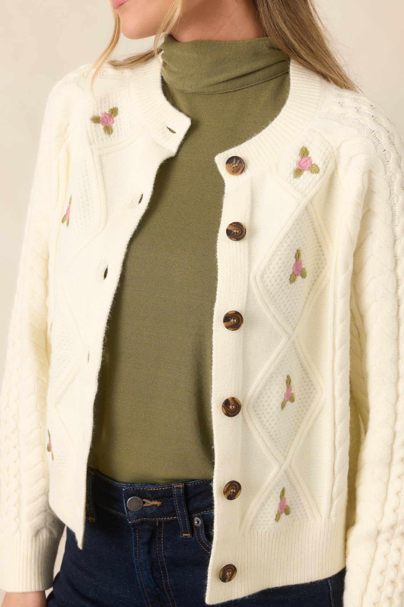 Close-up highlights the cable-knit stitching, tiny pink floral embroidery, textured fabric, and tortoise-style button.