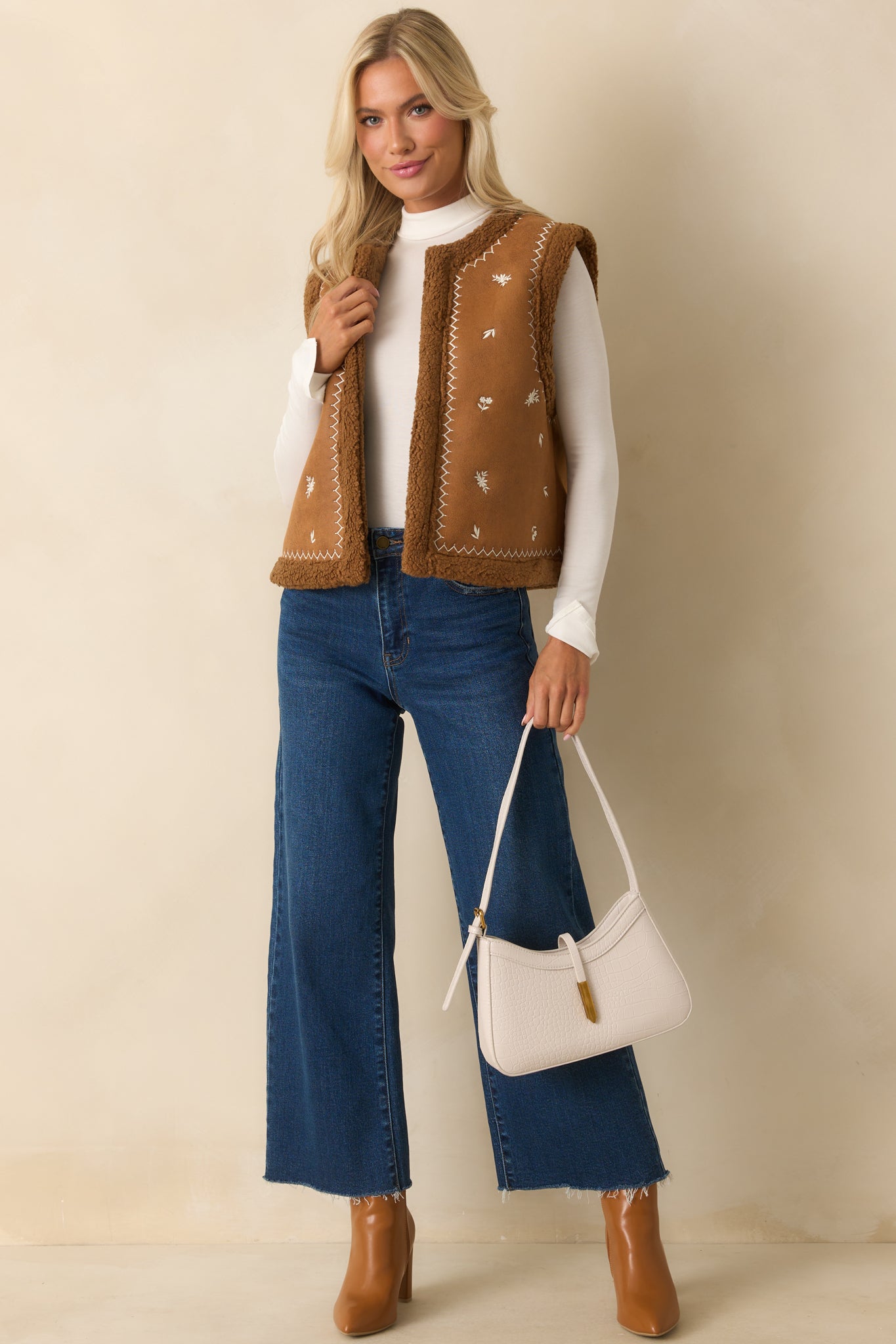 Caramel brown faux suede vest with open front, ivory floral embroidery, faux shearling trim, and straight hemline.
