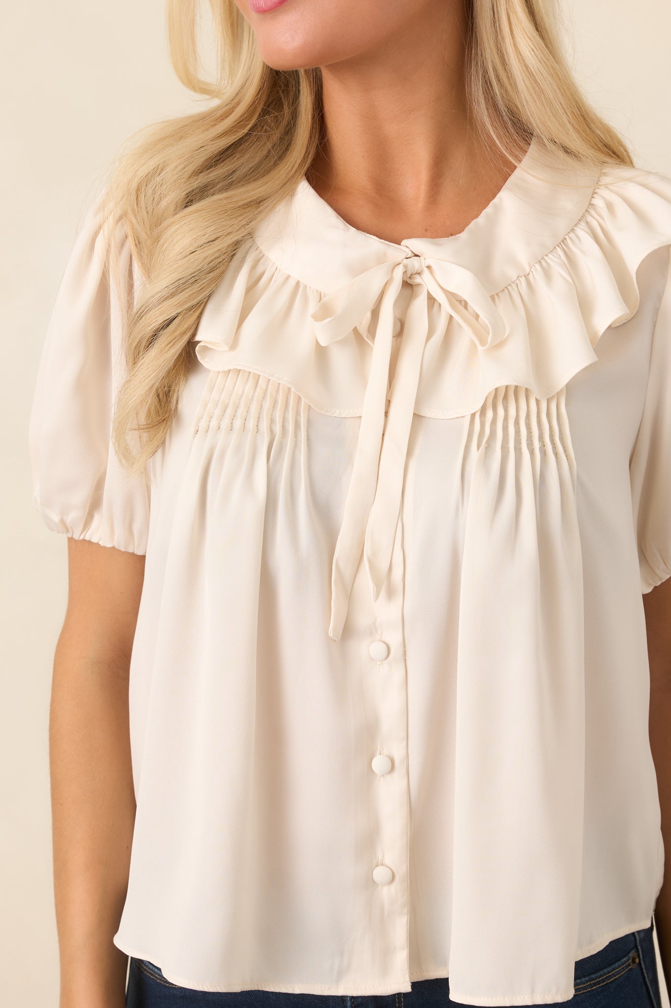 Close-up of the champagne top highlighting the wide ruffled collar, tie neckline, covered button detailing, and softly gathered fabric.