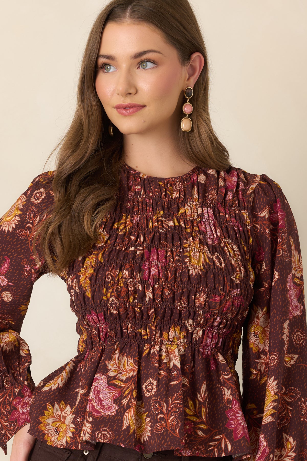 Smocked chocolate brown floral top with a round neckline, colorful botanical print, long puff sleeves, and softly flared peplum hem.